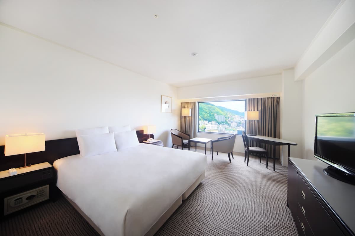 A hotel room with a large bed, a desk with a chair, a television, and a view of a cityscape through the window.
