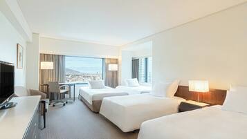 Quarto de hotel com duas camas, uma escrivaninha, uma cadeira e uma televisão. Há uma janela grande com vista para a cidade.