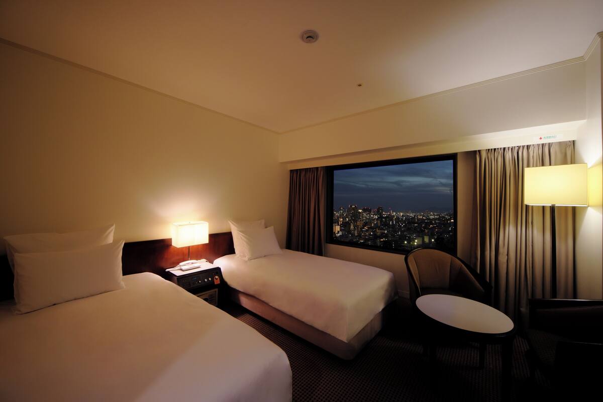 A hotel room with two beds, a desk, a chair, and a large window with a city view.