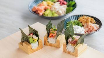 Tres rollos de sushi sobre bloques de madera, con alga marina por encima, un bol de sashimi variado al fondo y un bol de verduras encurtidas.