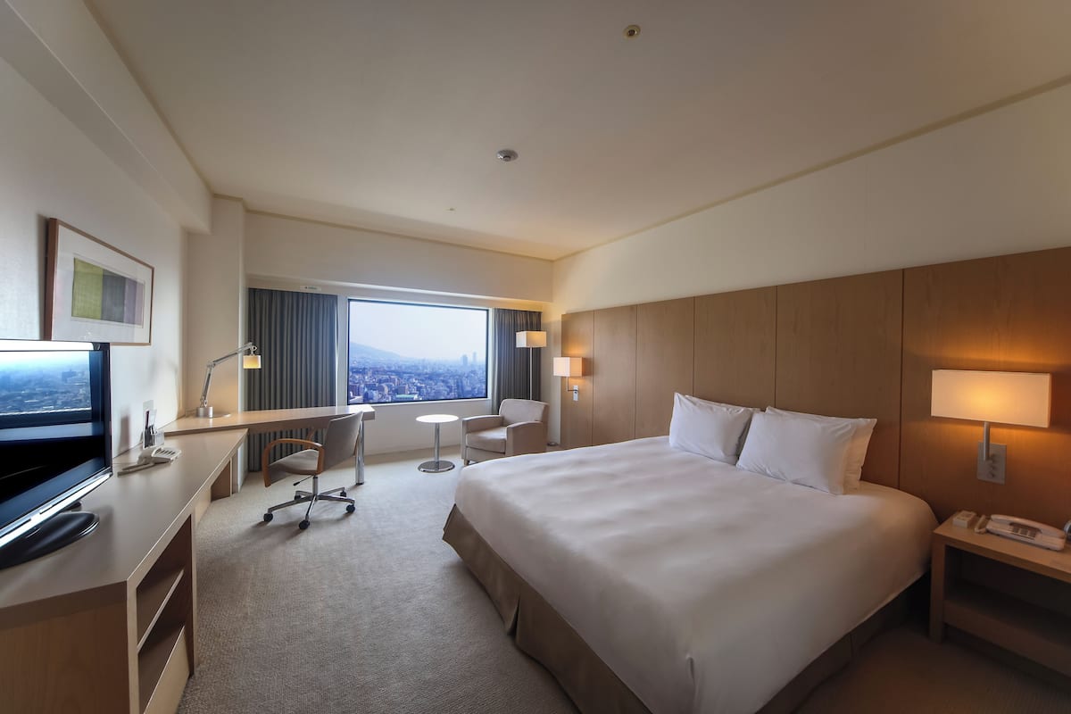 A hotel room with a large bed, a desk with a computer, a chair, a small table, and a TV.