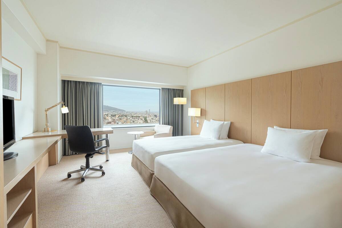 A hotel room with two beds, a desk, a chair, a TV, and a view of a cityscape.