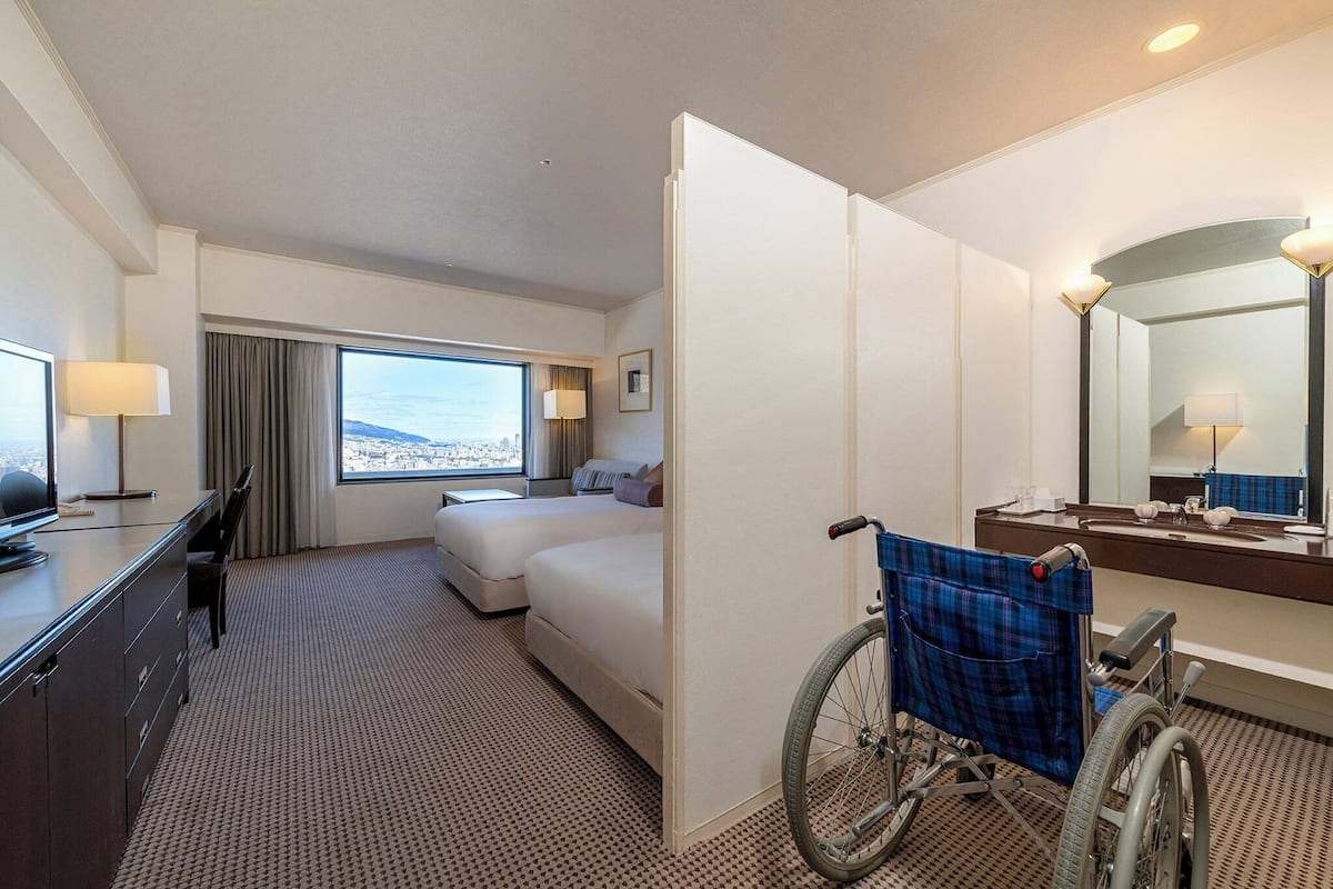 A hotel room with a large bed, a desk with a lamp, a wheelchair, and a bathroom with a mirror.