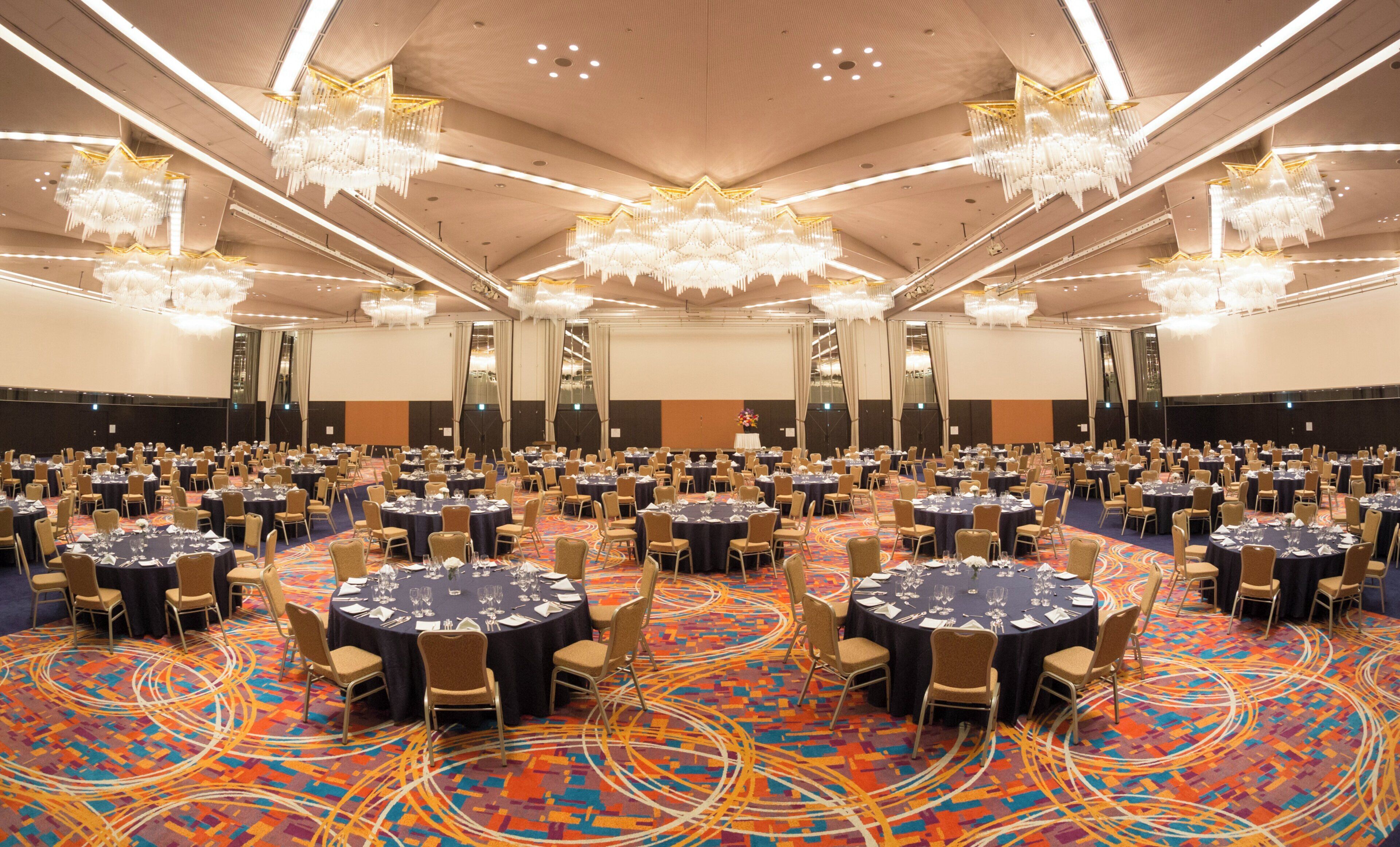 A large conference room with round tables and chairs, chandeliers, and a carpeted floor.