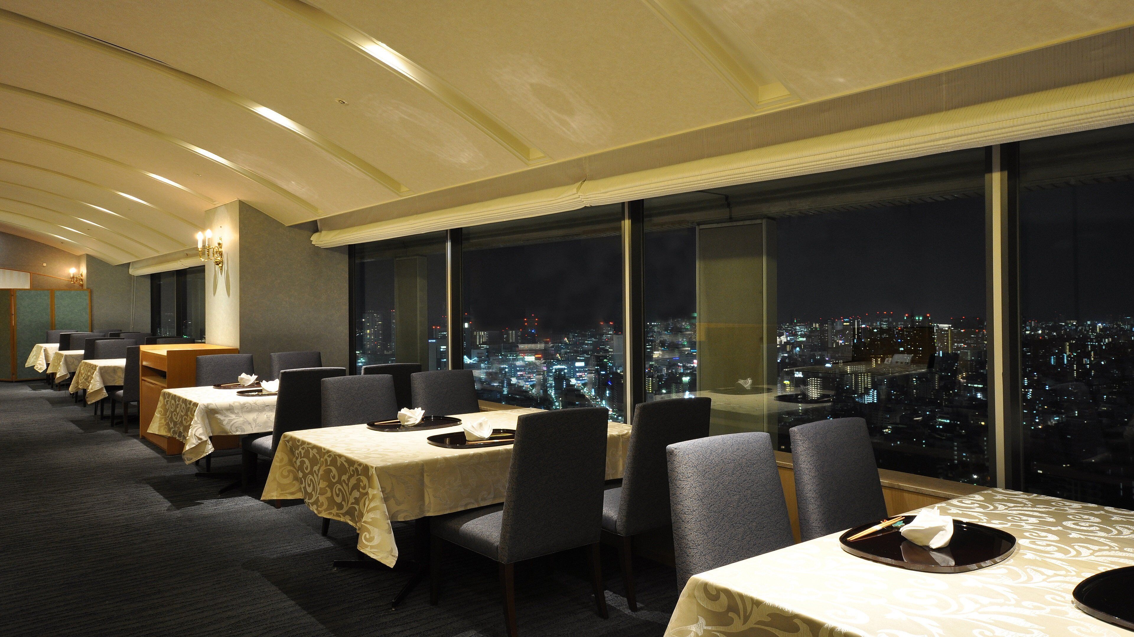 Un comedor de hotel con mesas dispuestas para los huéspedes, amplias ventanas que ofrecen vistas nocturnas de la ciudad.
