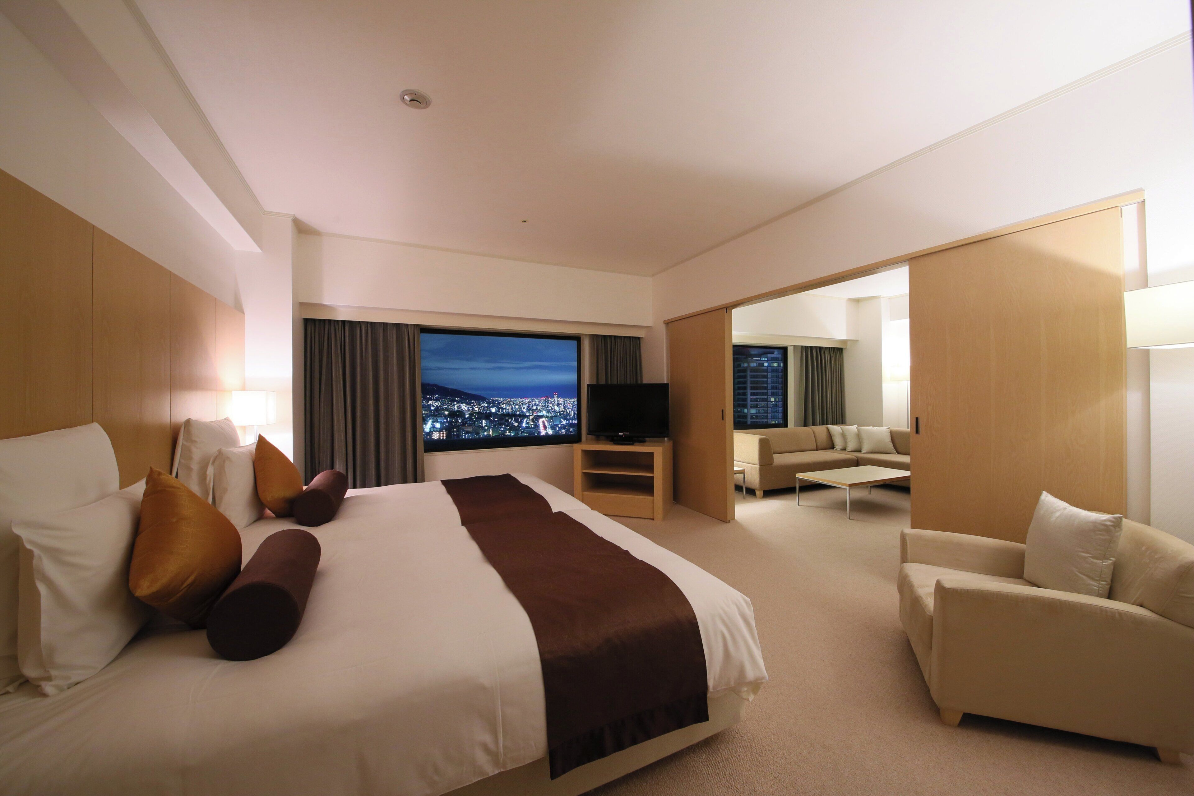 A hotel room with a large bed, a TV, a sofa, and a view of a cityscape.