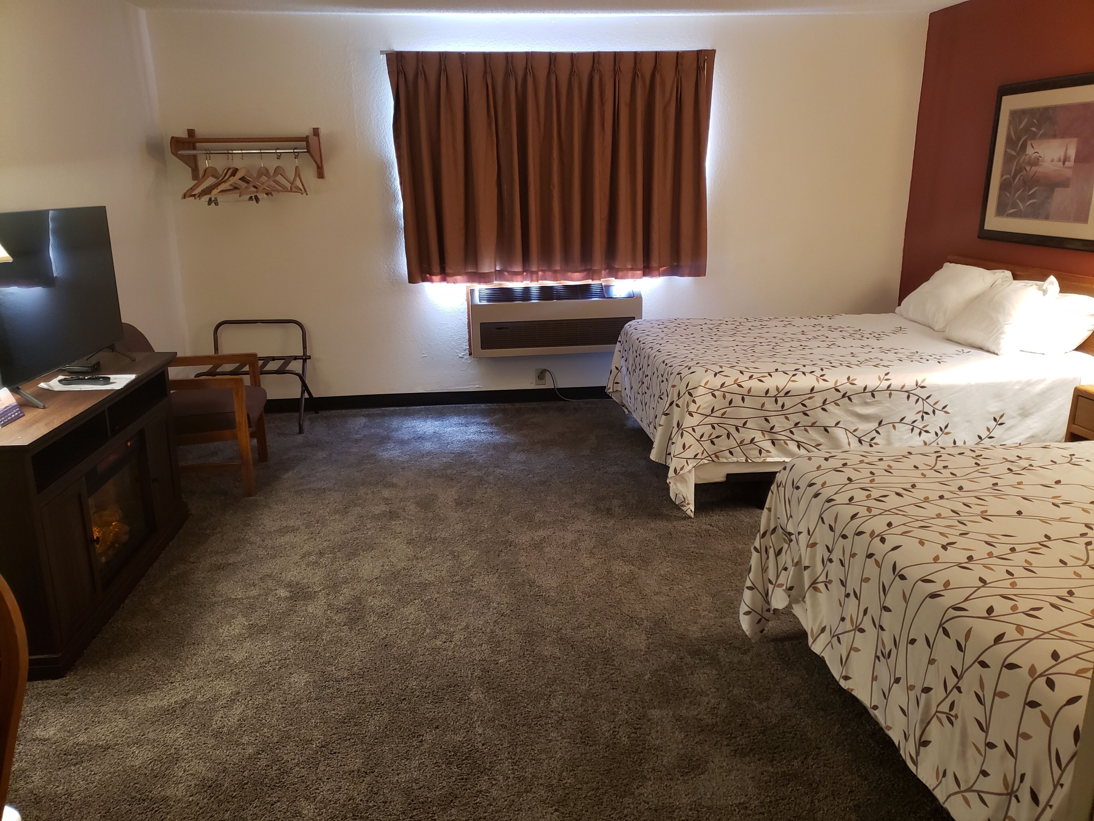 A hotel room with a TV, a chair, a bed with a leaf-patterned bedspread, a window with curtains, and a wall-mounted coat rack.