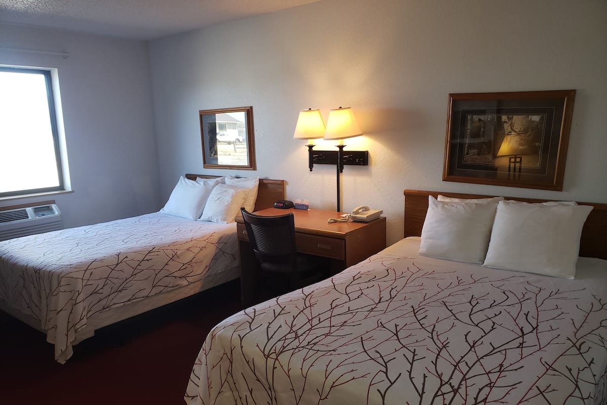 A hotel room with two beds, a desk, a chair, a lamp, a clock, and a framed picture on the wall.
