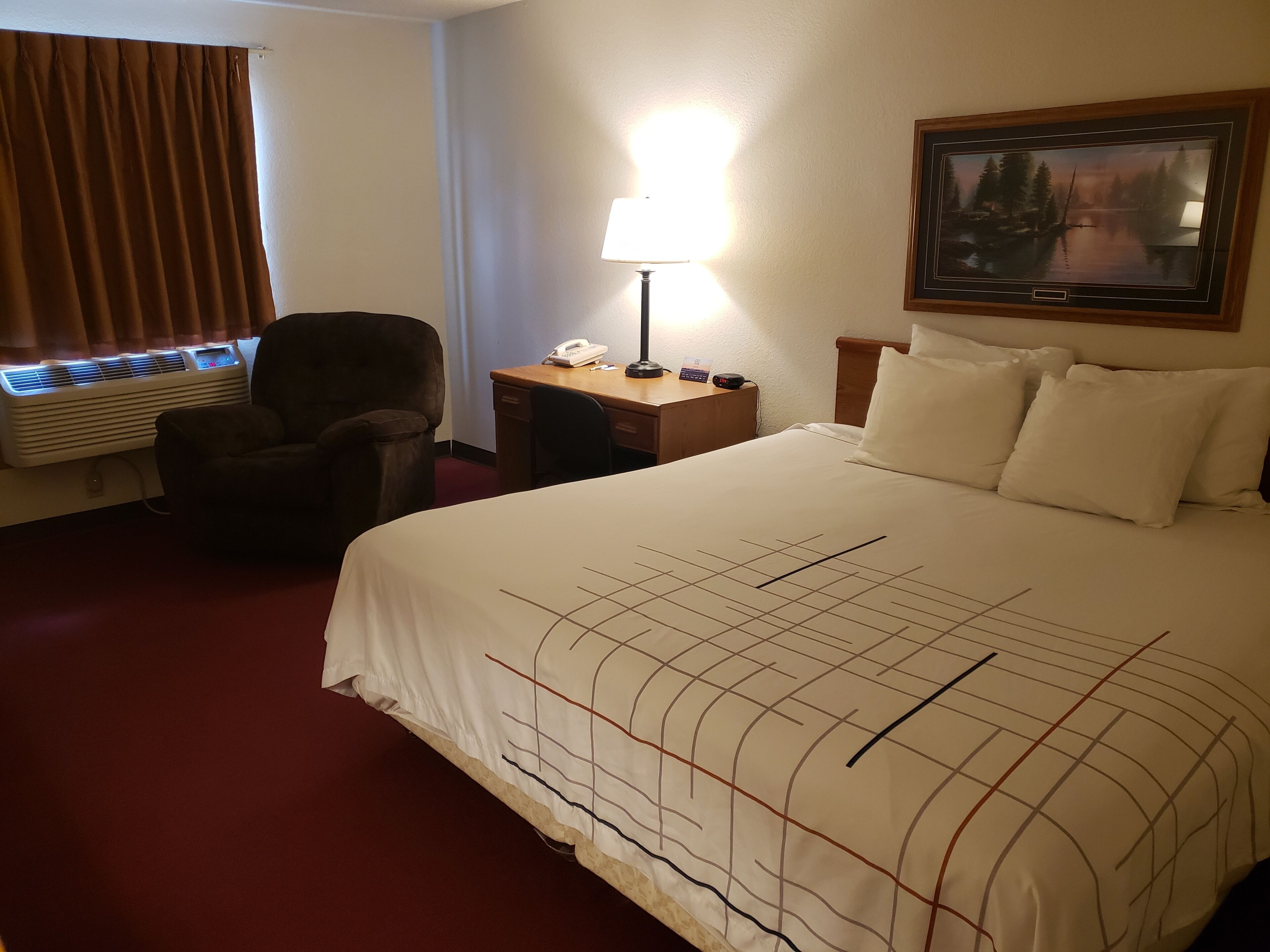 Habitación de hotel con cama, una silla, escritorio con lámpara, teléfono y un cuadro en la pared.