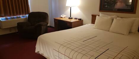Habitación de hotel con cama, una silla, escritorio con lámpara, teléfono y un cuadro en la pared.