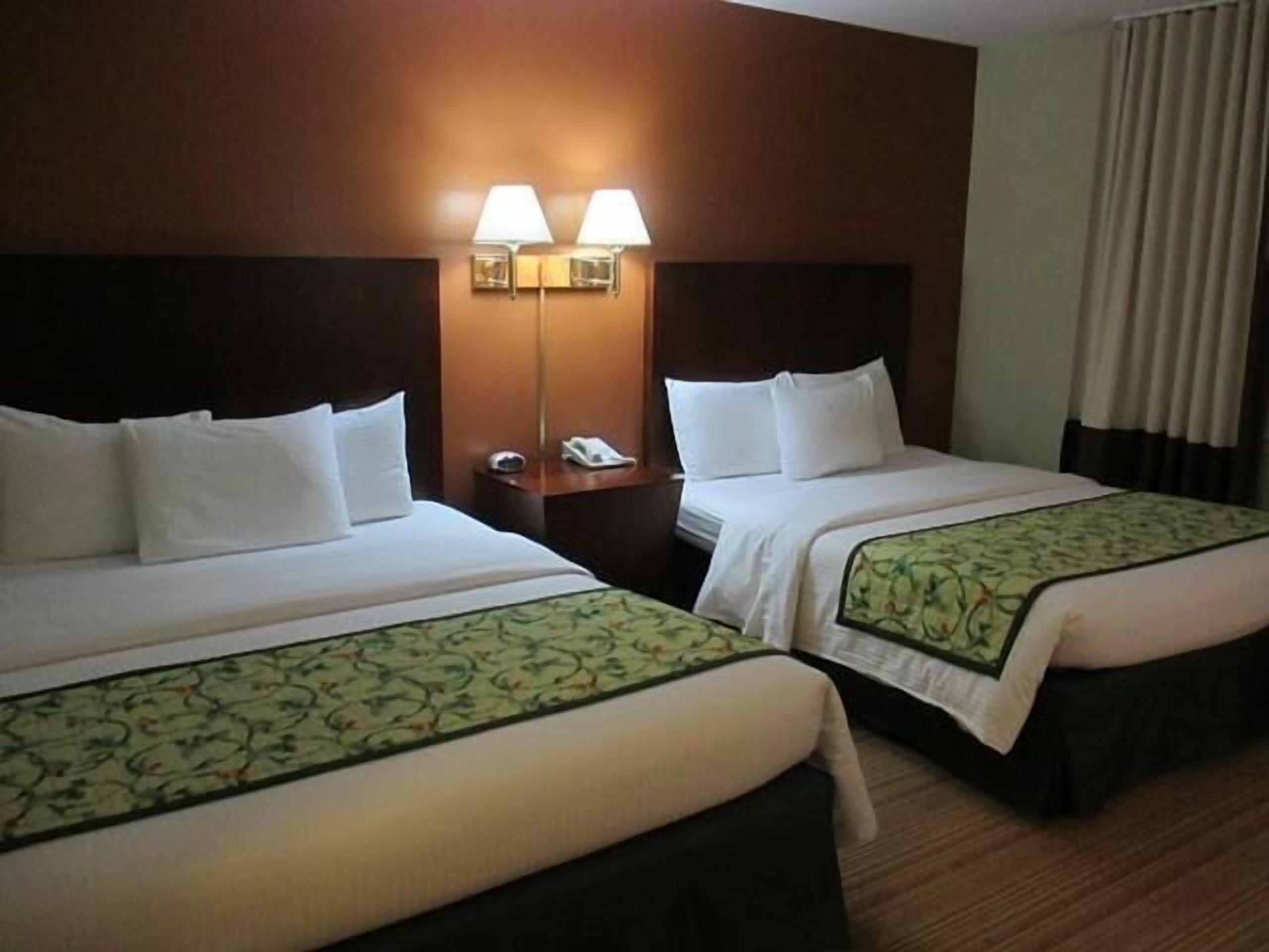 Quarto de hotel com duas camas, cada uma com roupa de cama branca e bordas decorativas verdes. Há luminárias fixadas na parede e um telefone na mesa de cabeceira entre as camas.