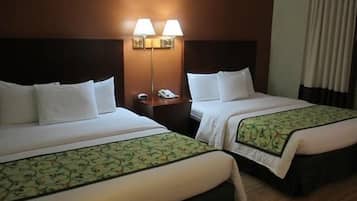 Quarto de hotel com duas camas, cada uma com roupa de cama branca e bordas decorativas verdes. Há luminárias fixadas na parede e um telefone na mesa de cabeceira entre as camas.