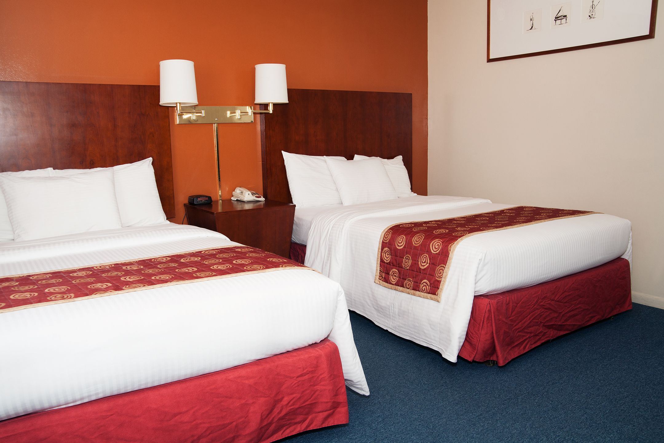 Hotellihuone, jossa on kaksi sänkyä. Jokaisessa sängyssä on valkoiset vuodevaatteet ja punaiset yksityiskohdat. Sängyn päädyssä on puinen koristepaneeli, yöpöydällä on lamppu ja seinällä on kehystetty kuva.