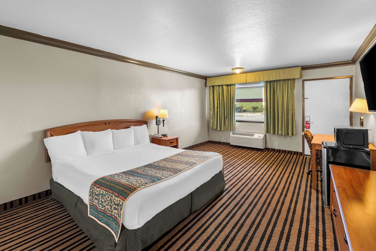 A hotel room with a large bed, a desk with a television, a window with curtains, and an air conditioning unit.