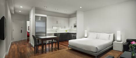 A modern hotel room with a large bed, a dining area, a kitchen, and a living area.