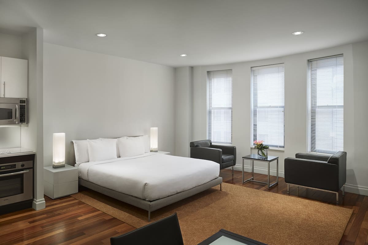 A modern hotel room with a large bed, a desk, a chair, and a small table. There is a microwave and a coffee maker on the left side. The room has large windows with blinds.