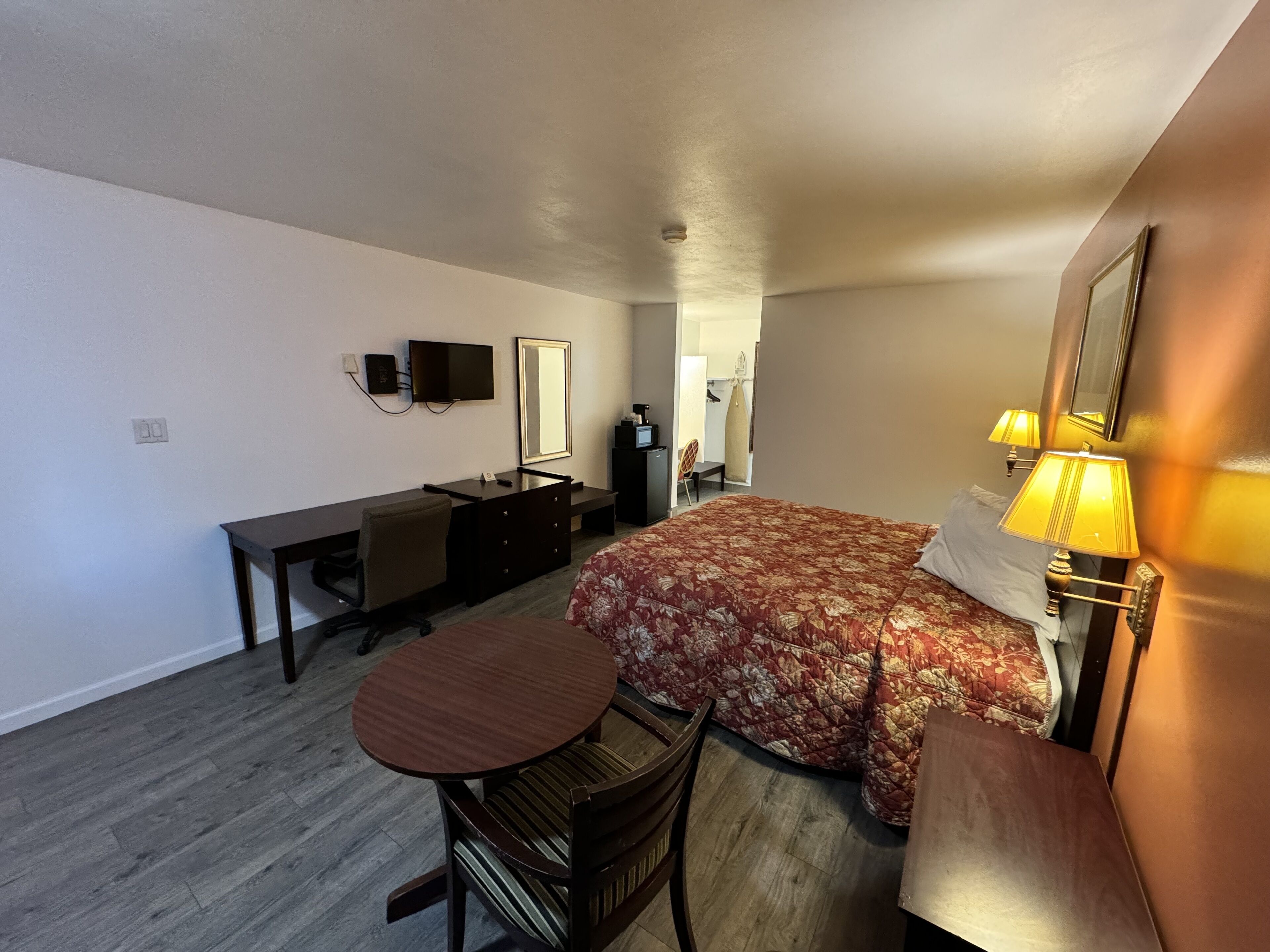 A compact hotel room with a bed, a desk, a chair, a small table, a TV, and a mirror.