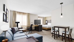 Una habitación de hotel moderna con sofá, escritorio, televisión, mesa de comedor con sillas y lámpara colgante.