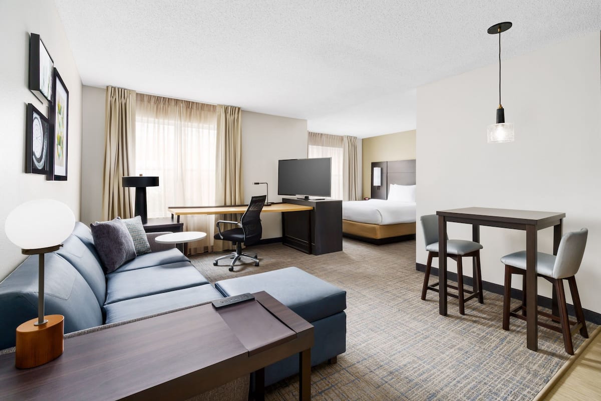 A modern hotel room with a sofa, a desk, a TV, a dining table with chairs, and a hanging light fixture.