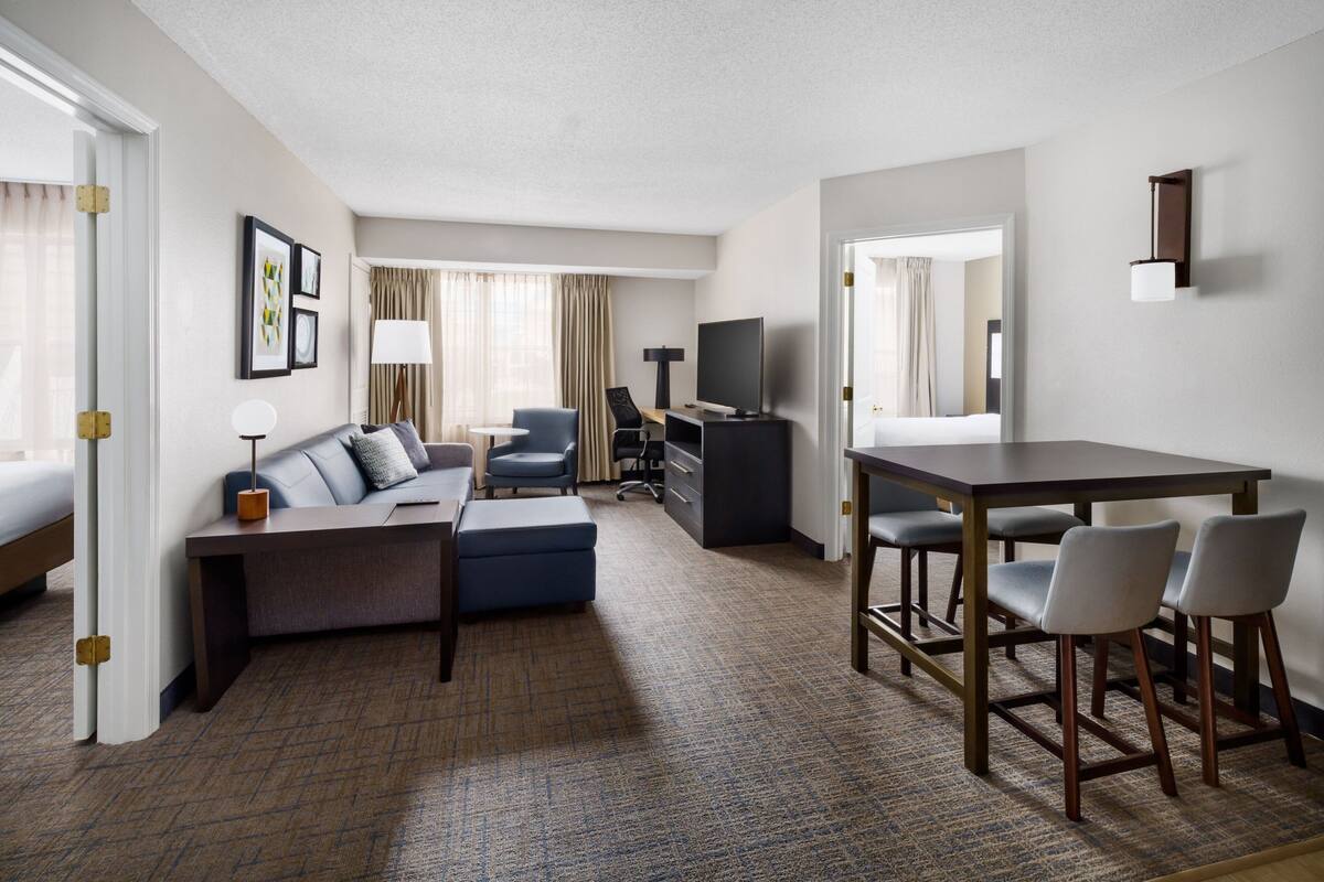 A modern hotel room with a sofa, a desk, a TV, and a dining area.