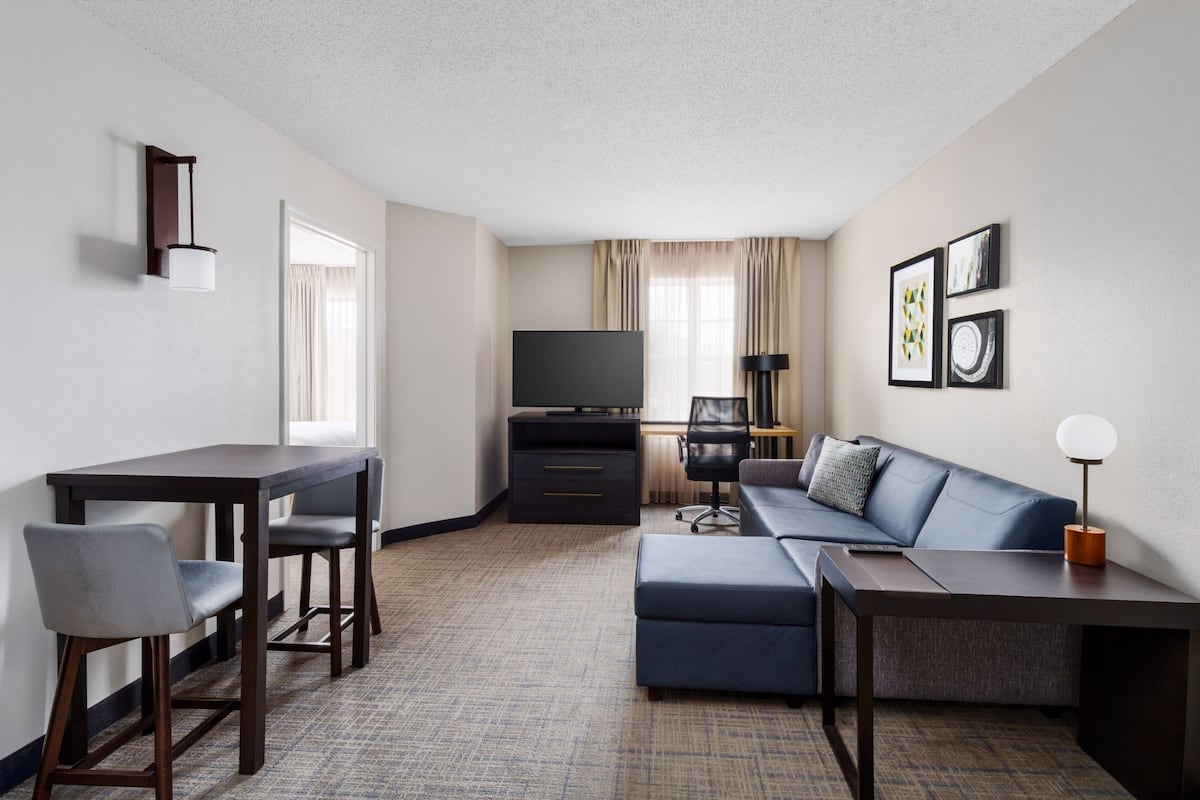 A modern hotel room with a dark wood desk, two chairs, a sofa, a TV, and a lamp.