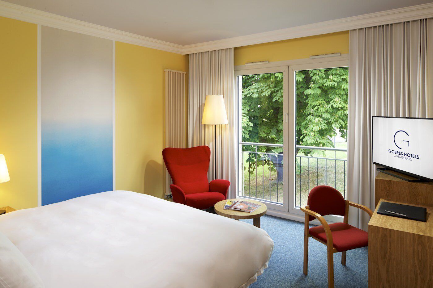 A hotel room with a bed, a red armchair, a smaller red chair, a small table with a magazine, and a TV displaying the hotel name.
