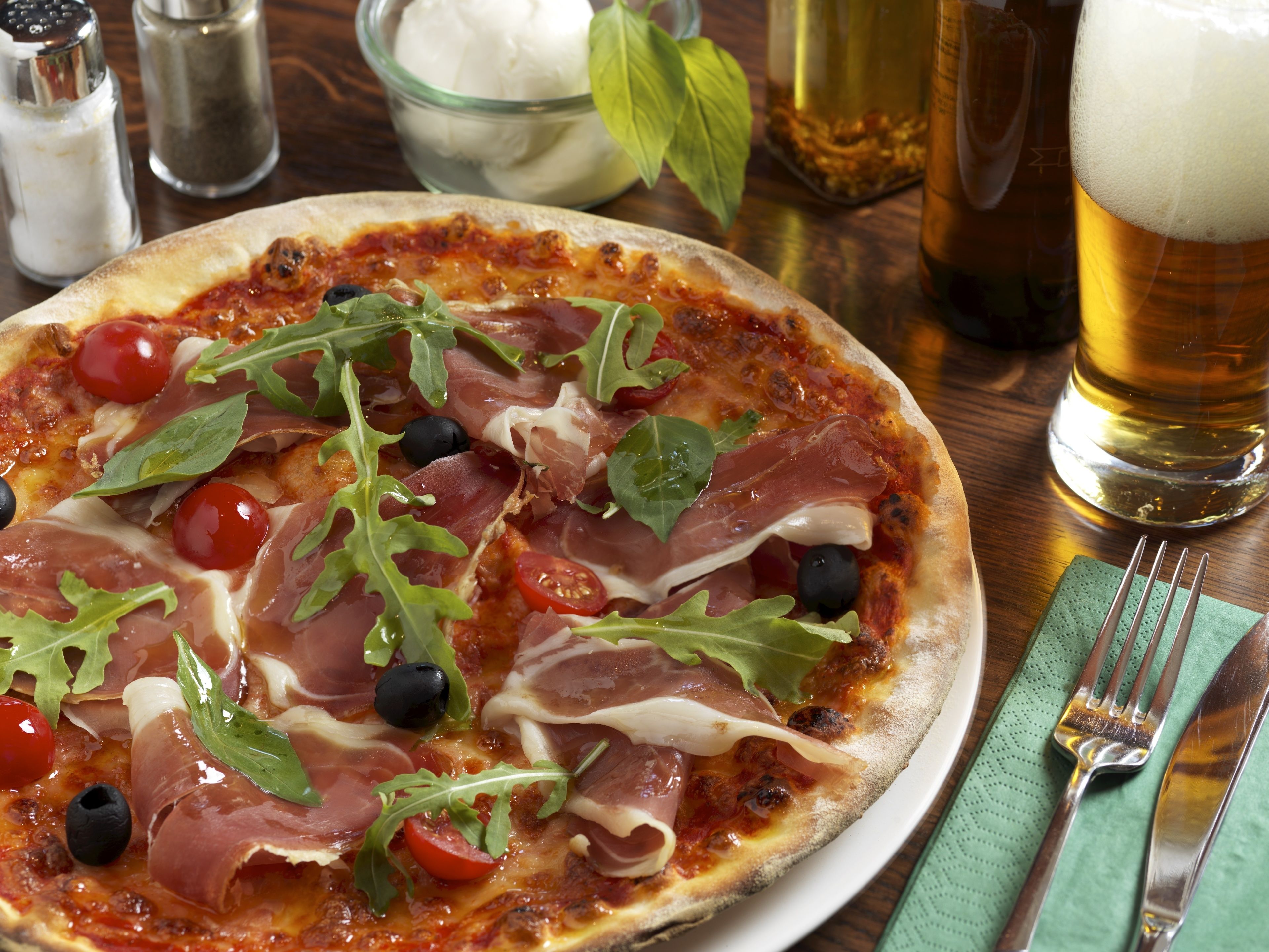 Une pizza garnie, un verre de bière et une serviette verte avec des couverts sur une table en bois.