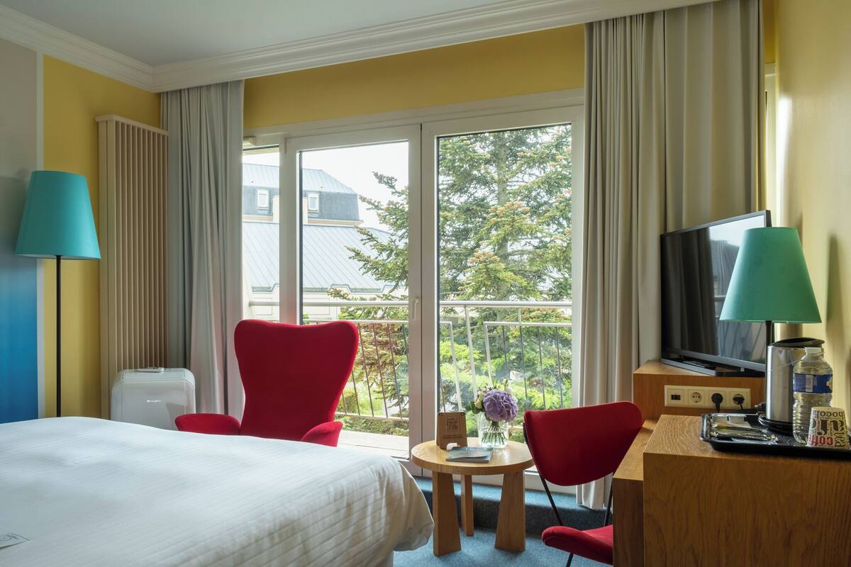 Habitación de hotel con cama, dos sillas rojas, un escritorio con televisor, una mesita con un florero y vistas a árboles y edificios a través del ventanal.
