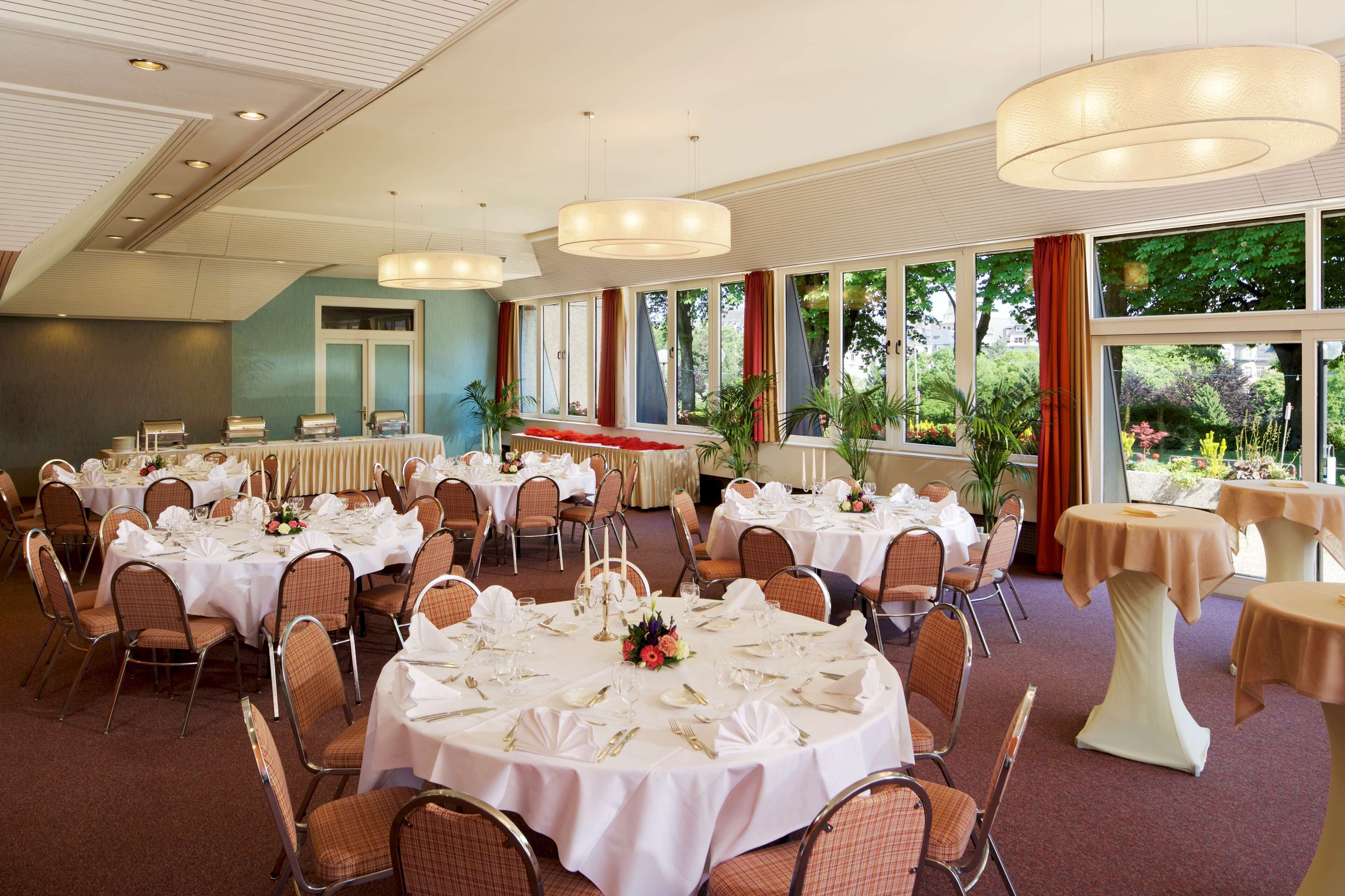 Un salón de banquetes con mesas redondas dispuestas para comer, rodeado de sillas y grandes ventanales que dan a vistas de vegetación.