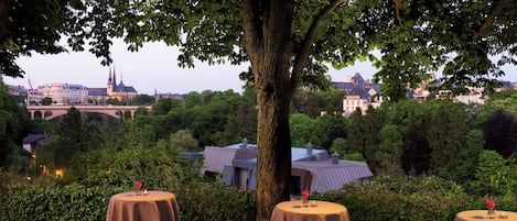Une vue sur un paysage urbain, avec un arbre au premier plan, des tables recouvertes de nappes et décorées de bougies, et un pont en arrière-plan.