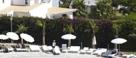 A pool area with lounge chairs and umbrellas, a clear view of the sea, and a building with white walls and a tiled roof.