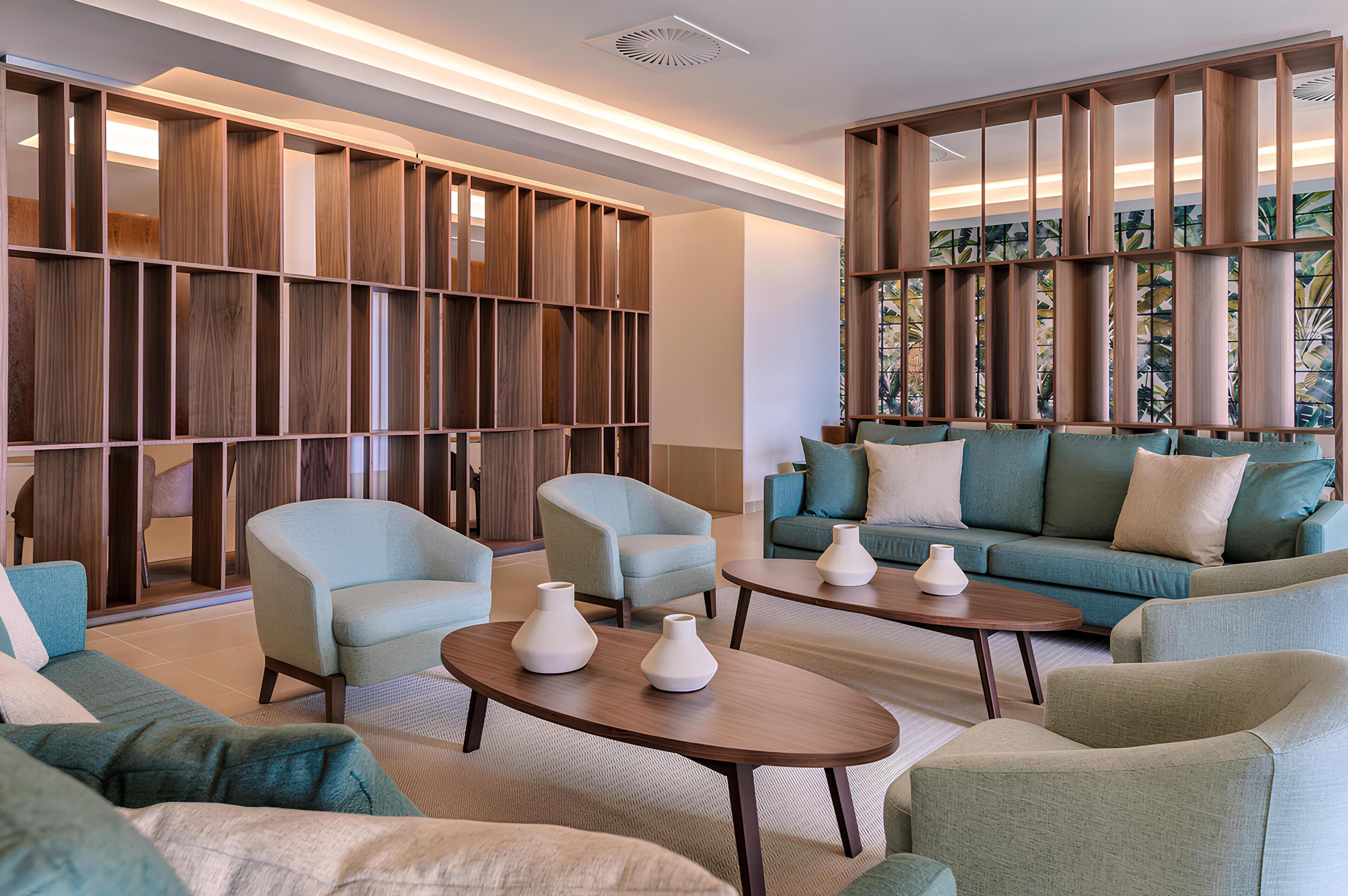 A modern living room with a sectional sofa, armchairs, and a coffee table. There is a wooden bookshelf and a large window with a view.