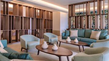 A modern living room with a sectional sofa, armchairs, and a coffee table. There is a wooden bookshelf and a large window with a view.