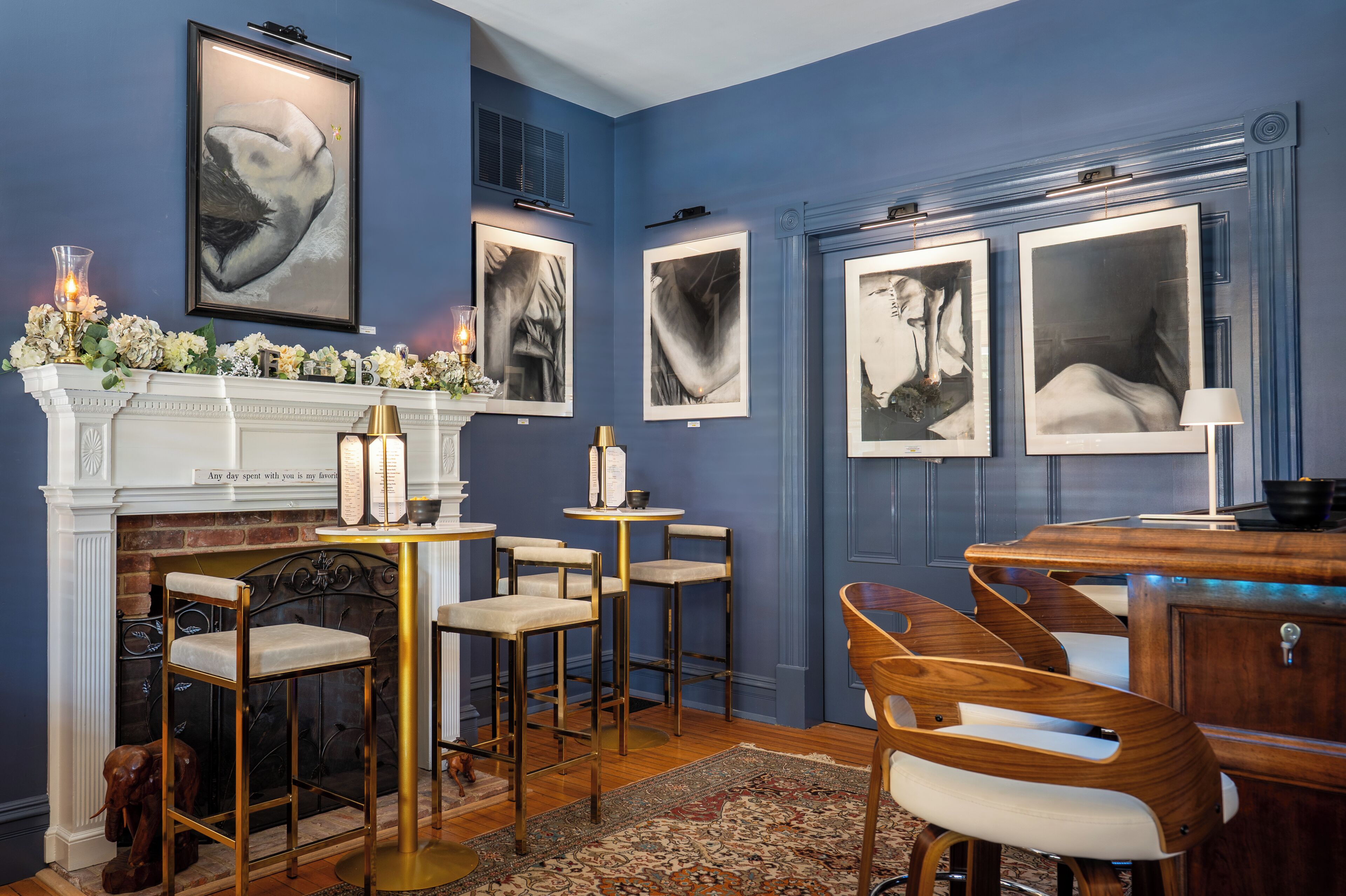A room with a blue wall, a fireplace, framed artwork, and a dining area with chairs and tables.