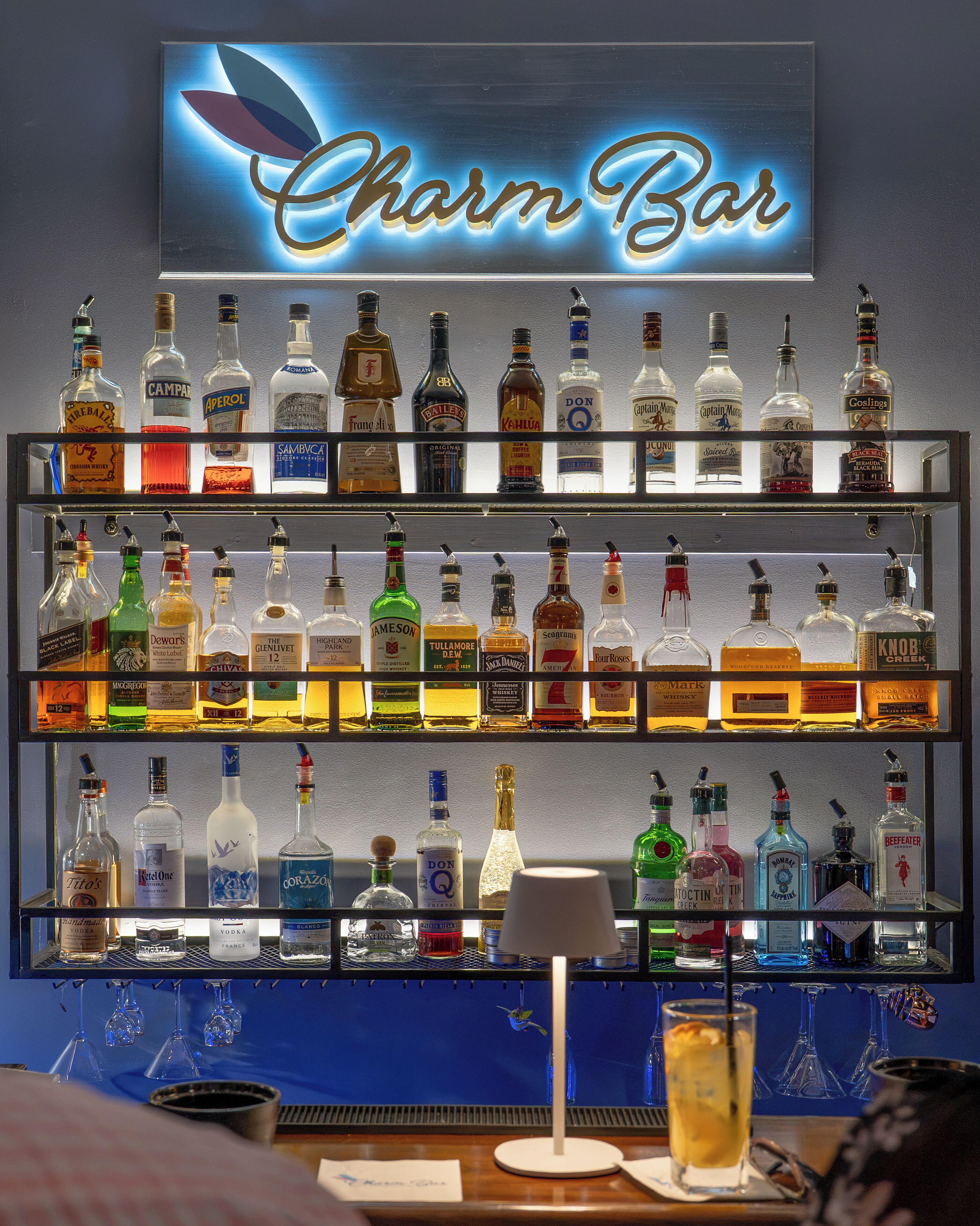 A bar with a neon sign reading 'Charm Bar' above a shelf of various liquor bottles.