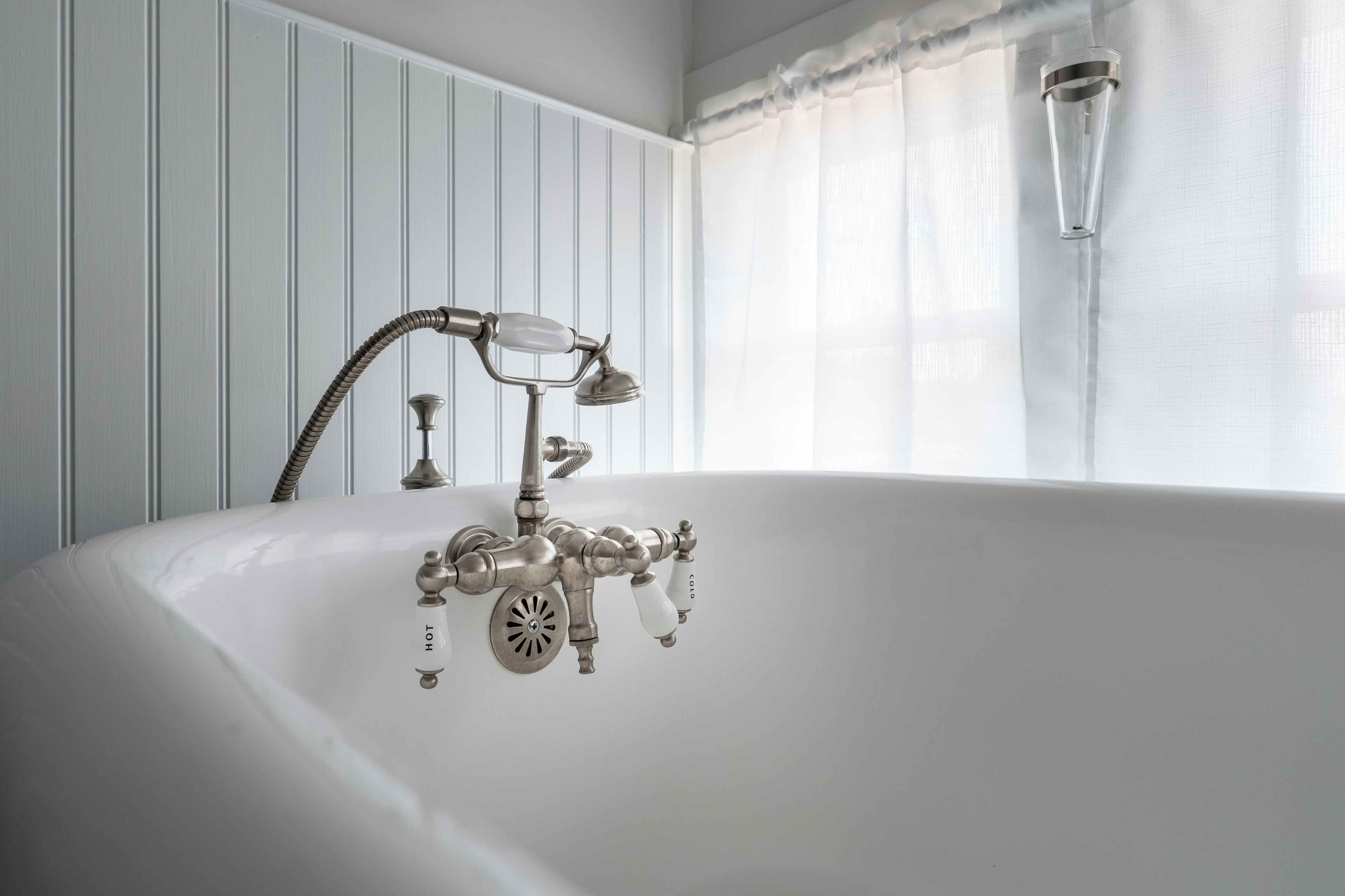 A classic white bathtub with a vintage faucet and showerhead, positioned in a bathroom with white paneled walls and a window with sheer curtains.