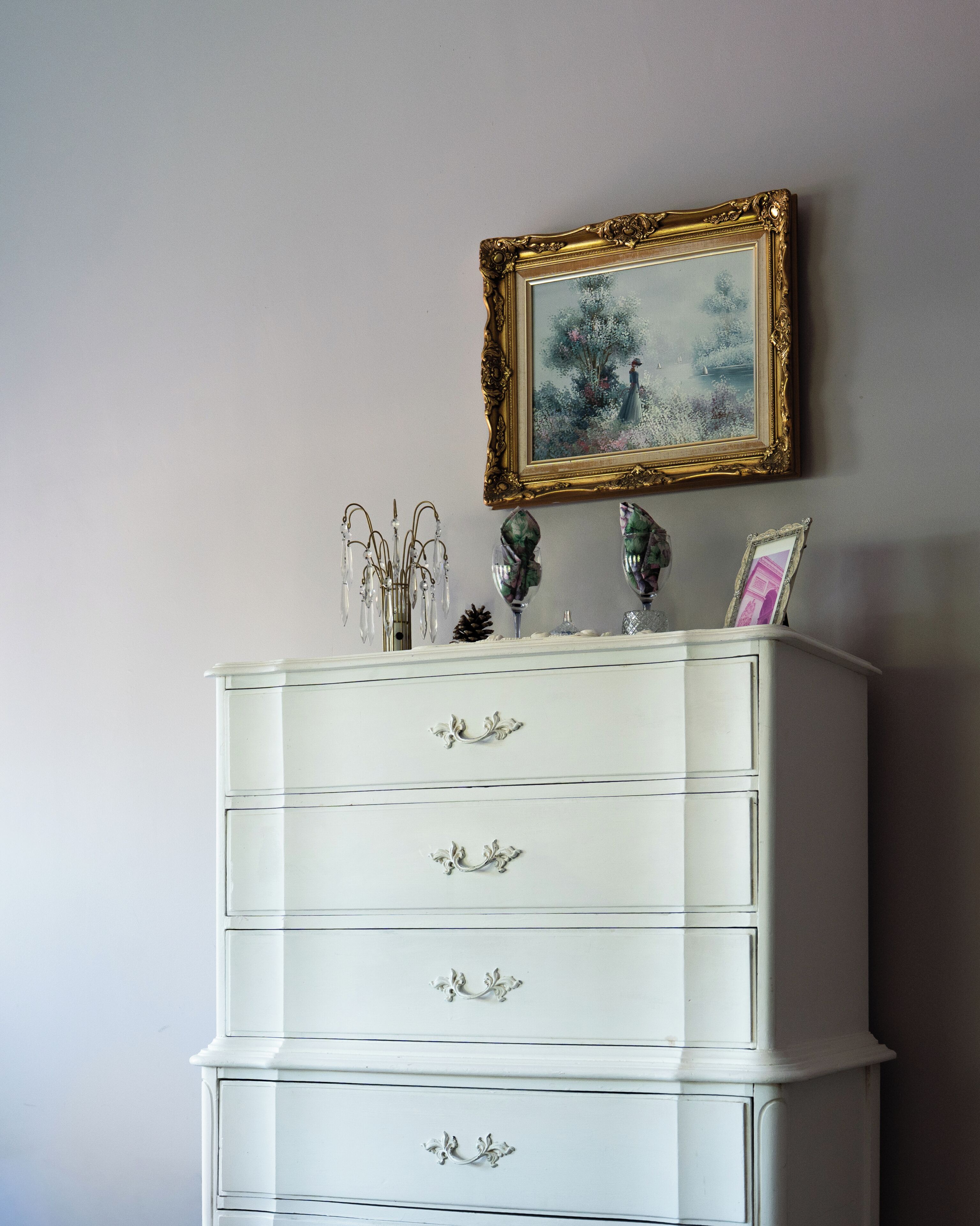 A white dresser with ornate handles and a gold-framed picture above it.
