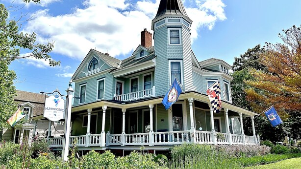 Una gran casa de estilo victoriano de dos pisos, con una torre destacada, numerosas ventanas y un porche.