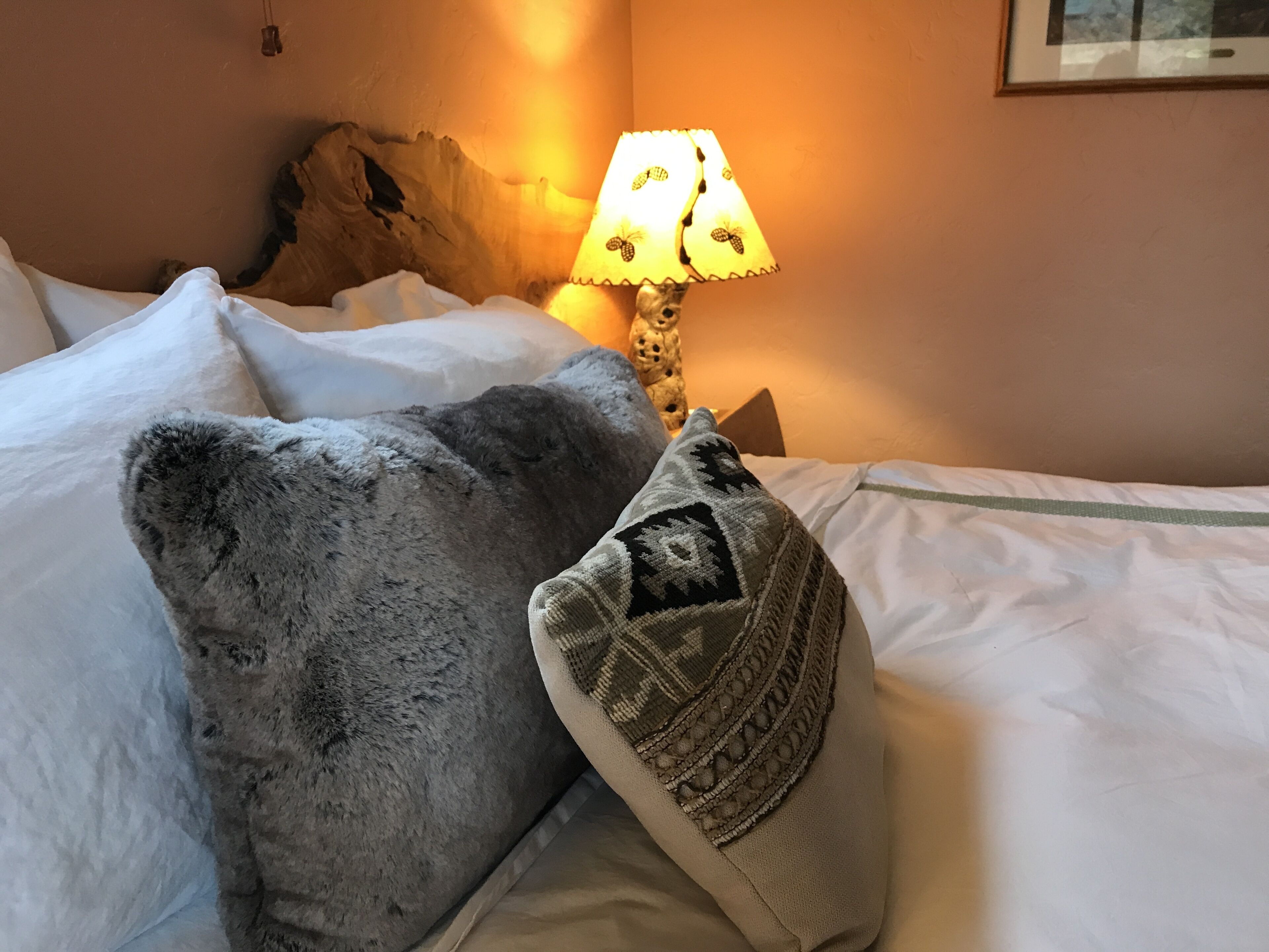 Un lit avec du linge de lit blanc, un oreiller moelleux de couleur grise et un autre oreiller à motifs. Une lampe avec un abat-jour à motifs est posée sur une table de chevet.