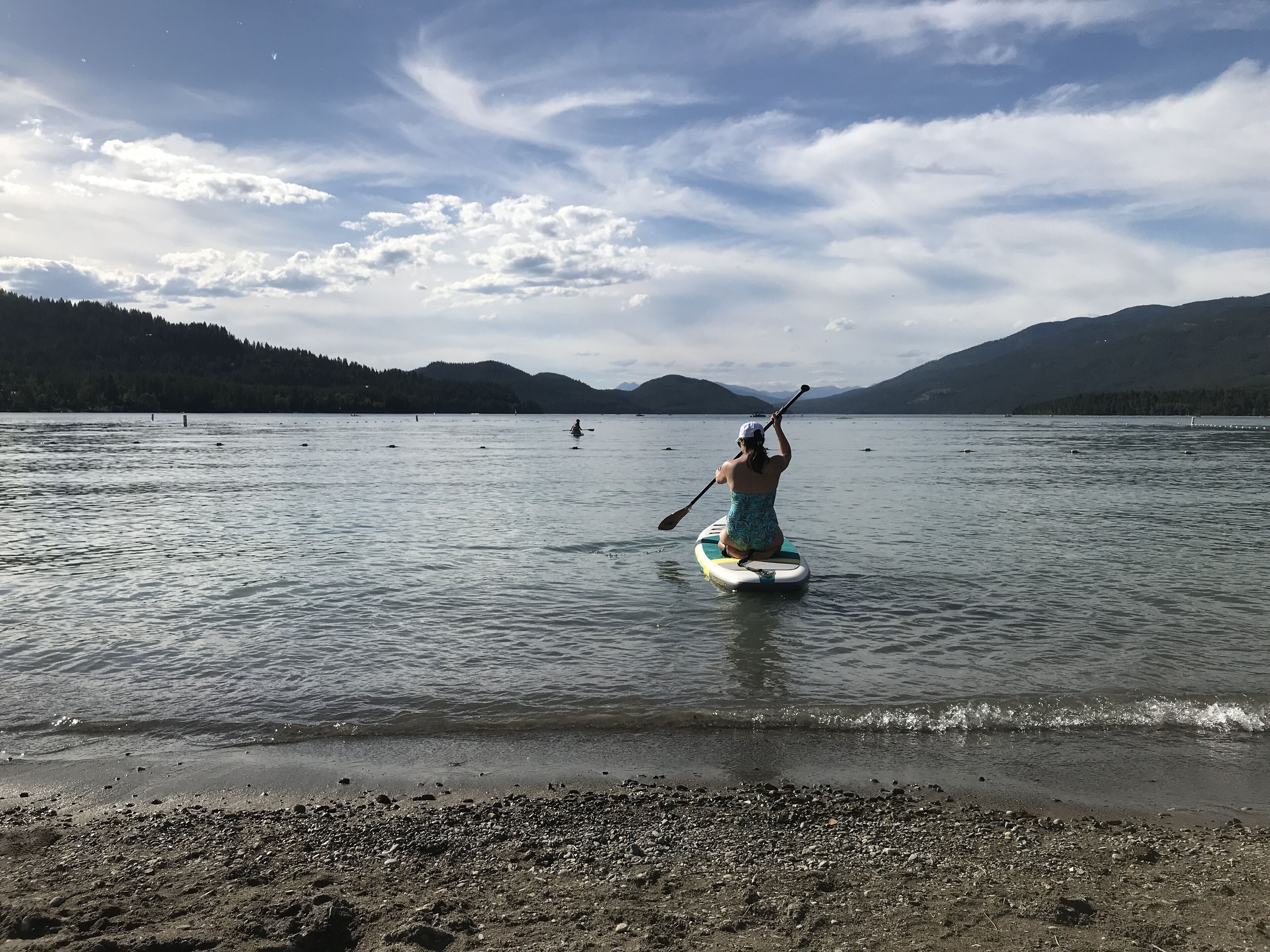 Eine Person steht auf einem Paddleboard auf einem ruhigen See, im Hintergrund Berge.