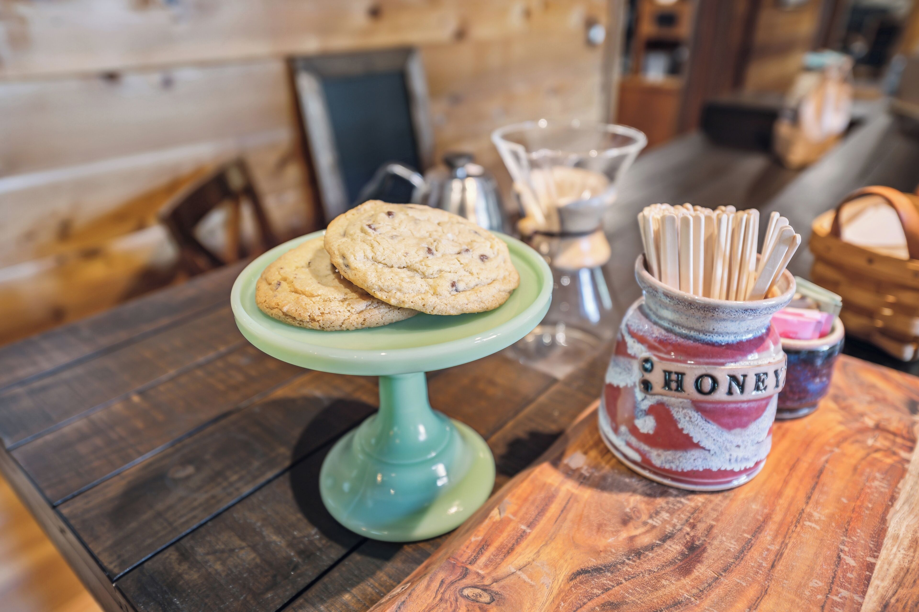 A wooden table with a green cake stand holding two cookies, a jar labeled "HONEY," and a container with wooden sticks.