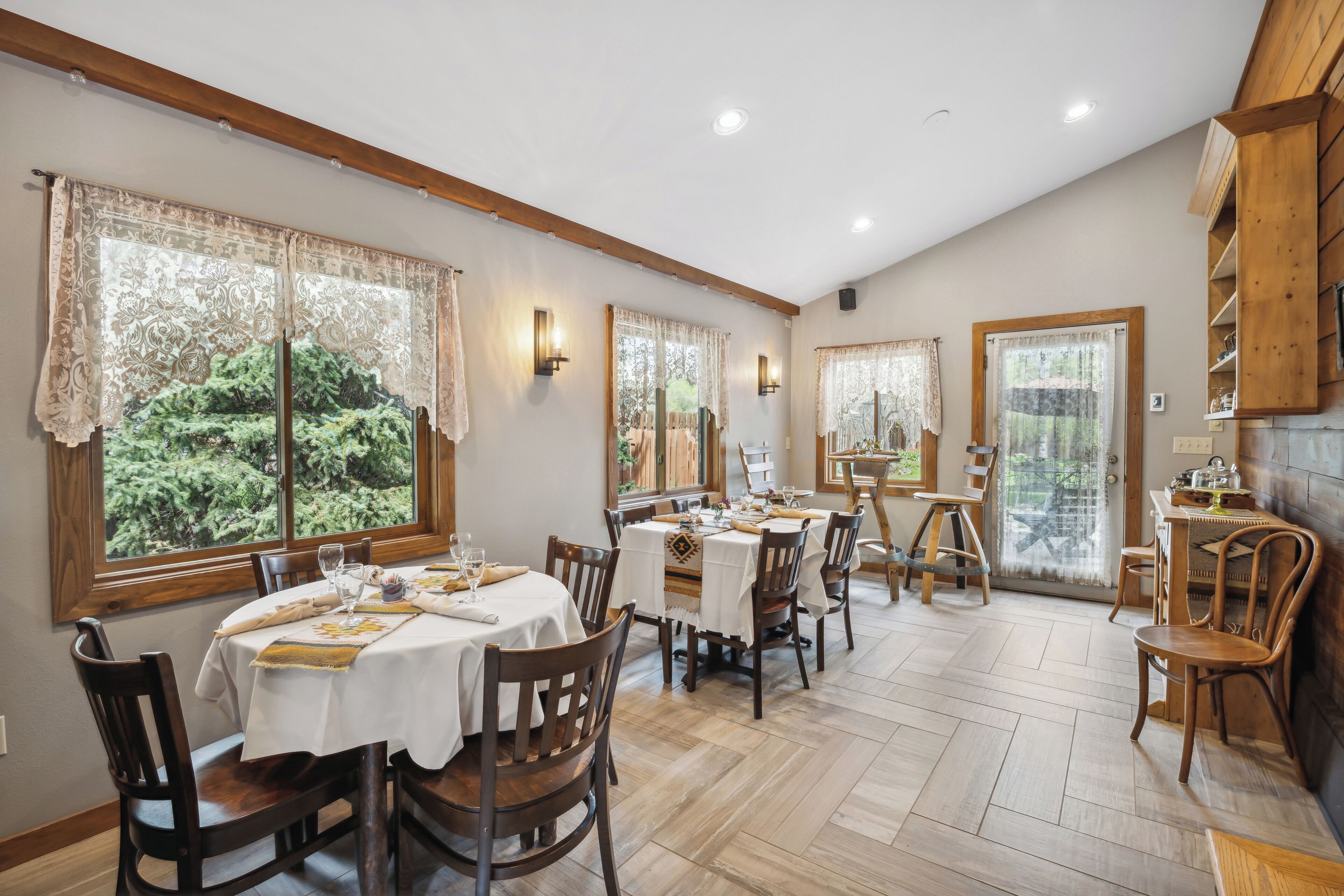 Uma sala de jantar com mesas e cadeiras redondas, janelas amplas com cortinas de renda e teto de madeira.