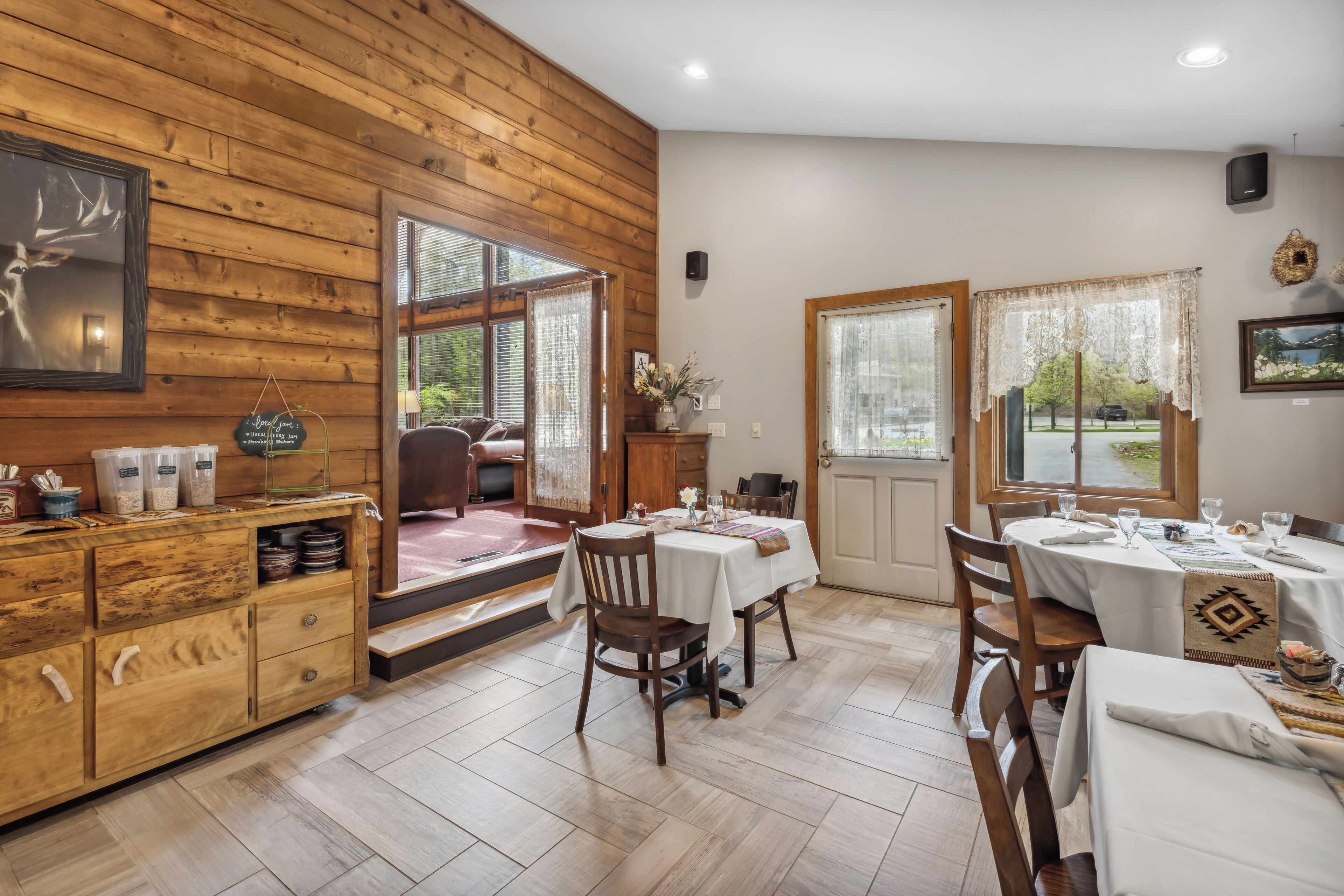 A dining area with wooden walls, white tables, and chairs. There are windows with curtains, a wooden cabinet with storage, and a door leading outside.