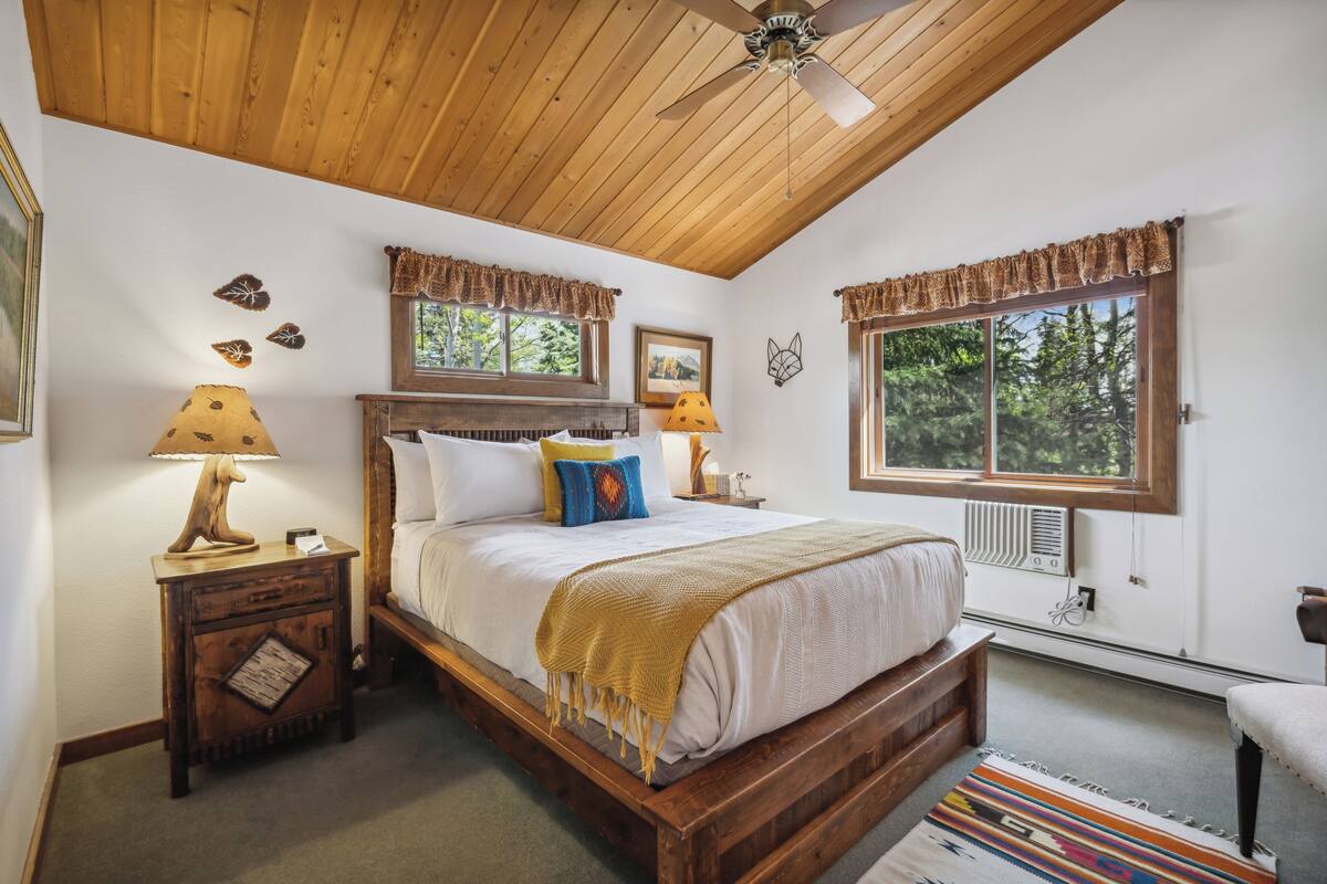 Un dormitorio con techo de madera, una cama con colcha blanca, dos mesitas de noche con lámparas y un ventanal con vistas a árboles.