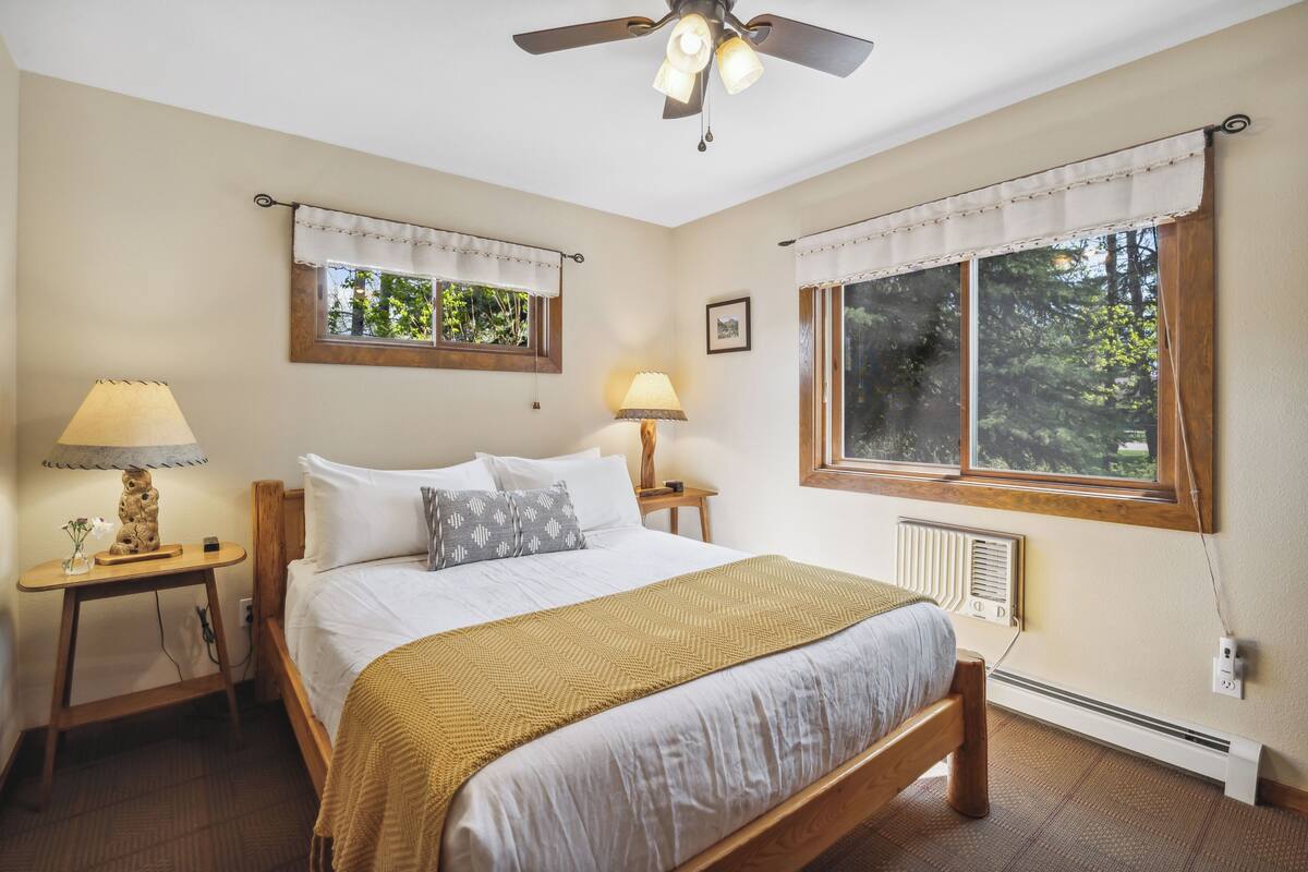 Un dormitorio con una cama, dos mesitas de noche, un ventilador de techo y dos ventanas con cortinas.