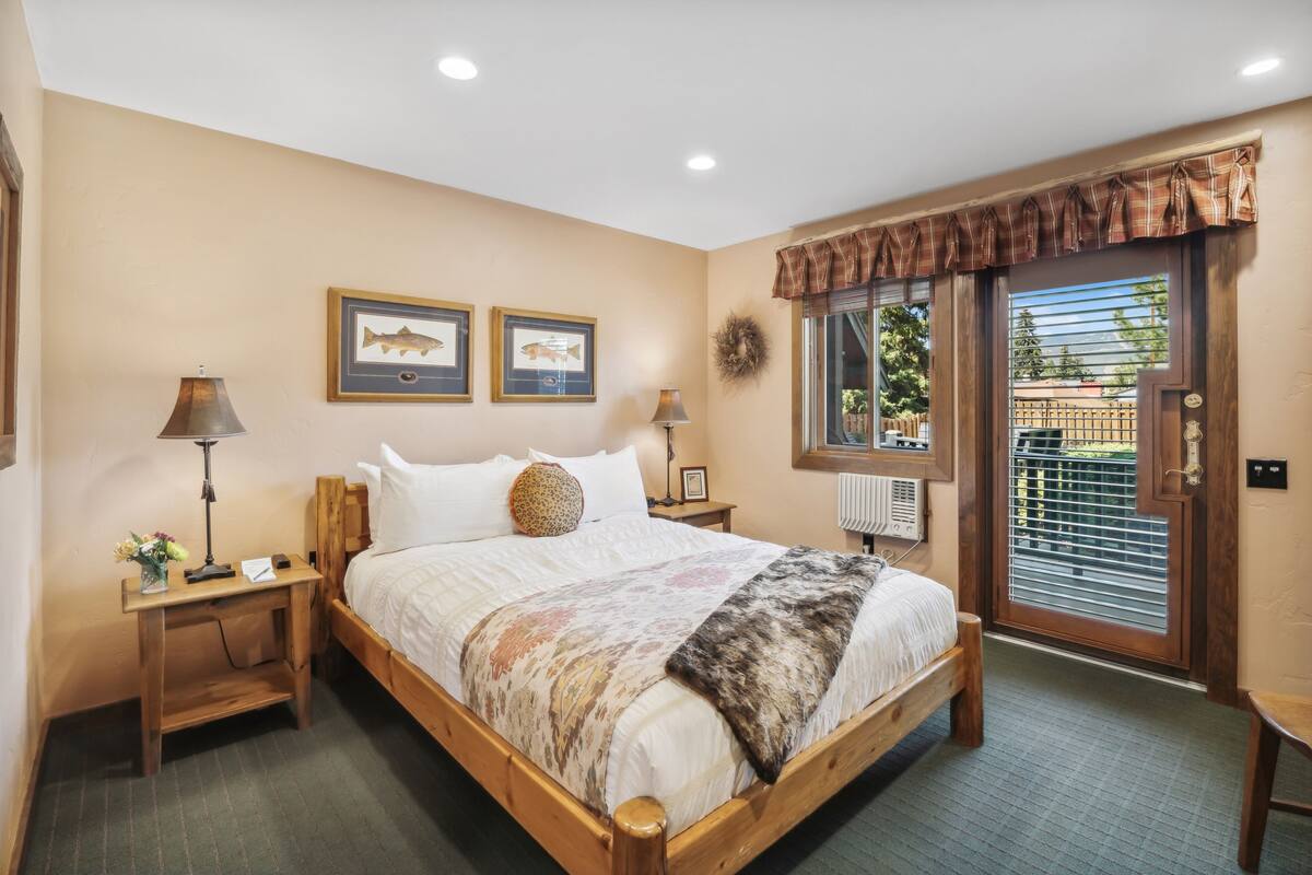 Un dormitorio con una cama de madera, dos mesitas de noche, una mesita auxiliar, una ventana con persianas y una puerta con cortina.