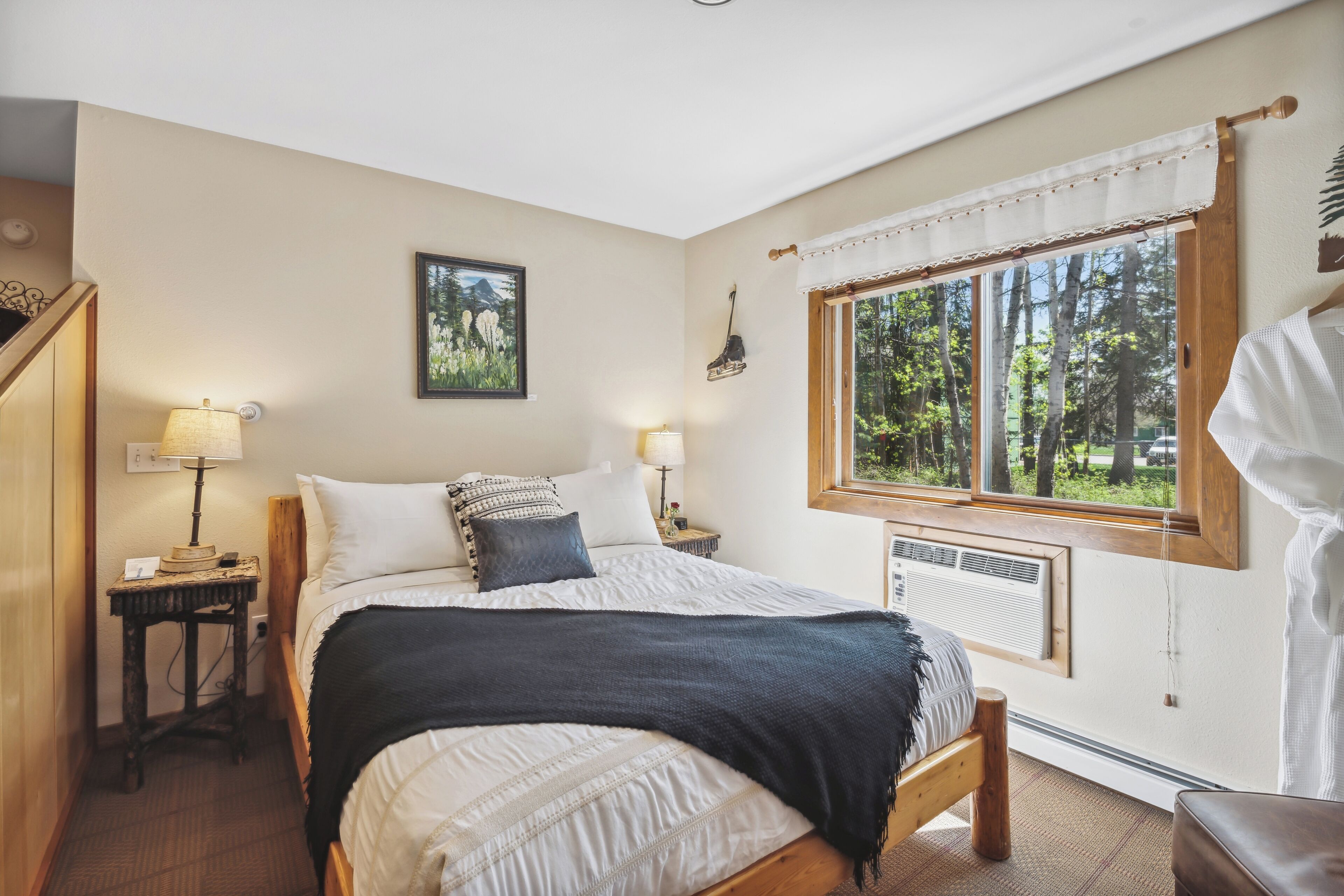 Un dormitorio con cama, dos mesitas de noche con lámparas, una ventana con vistas a árboles y un cuadro colgado en la pared.
