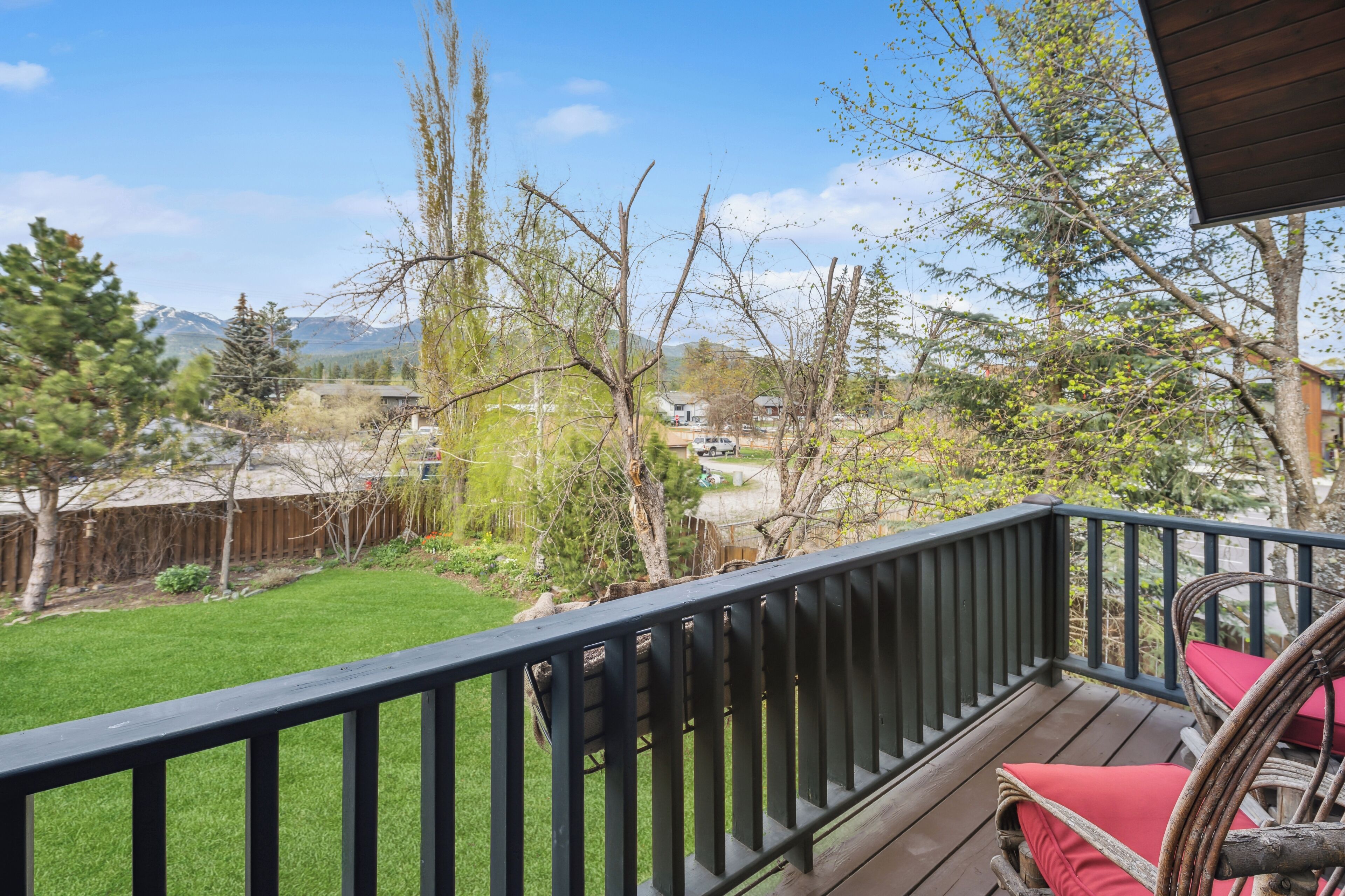 Une terrasse en bois avec une rambarde noire, une chaise en osier avec des coussins rouges, et une vue sur une pelouse verdoyante et des montagnes lointaines.