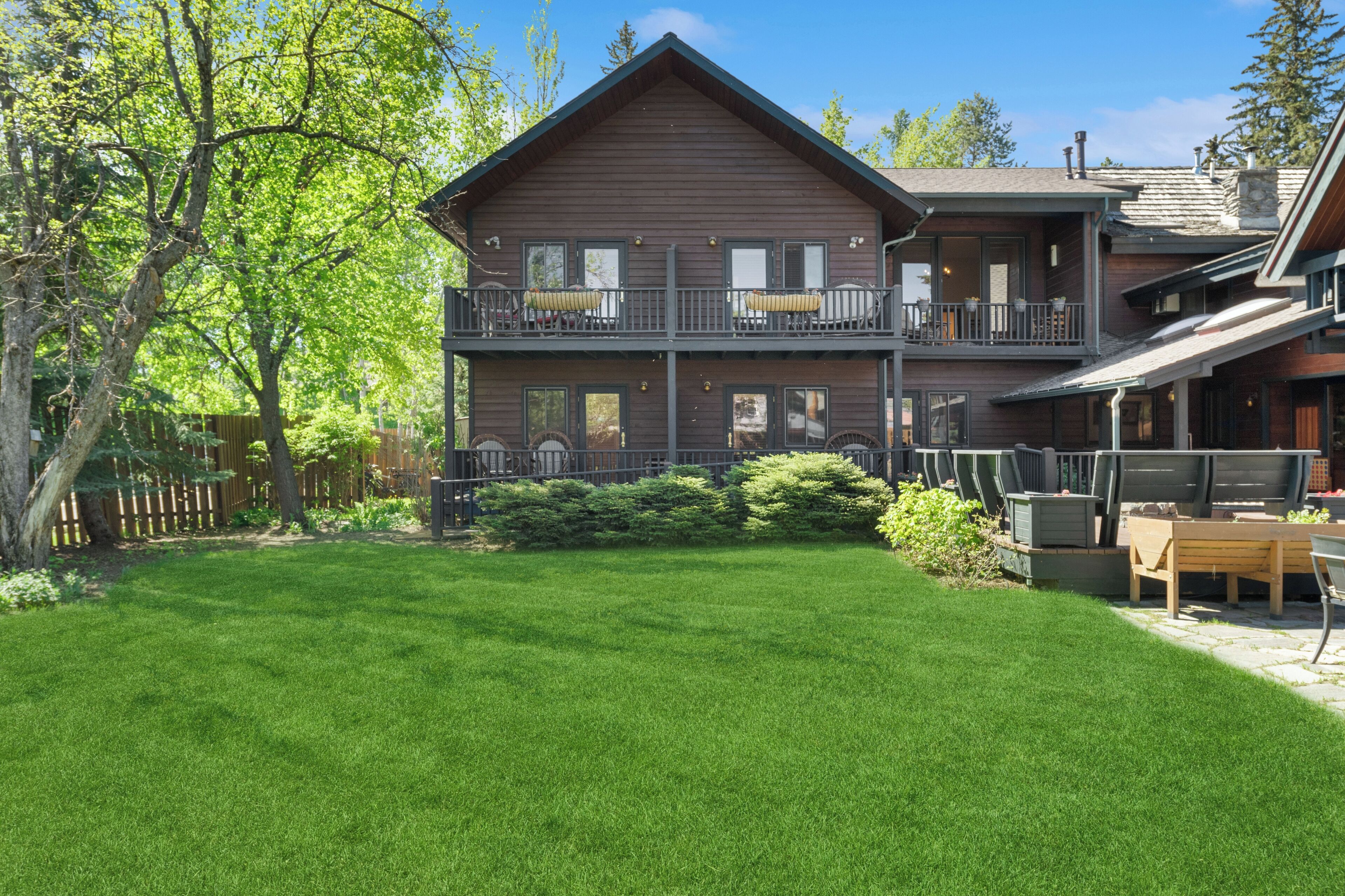 A two-story wooden house with a balcony, a well-maintained lawn, and a patio area with outdoor furniture.