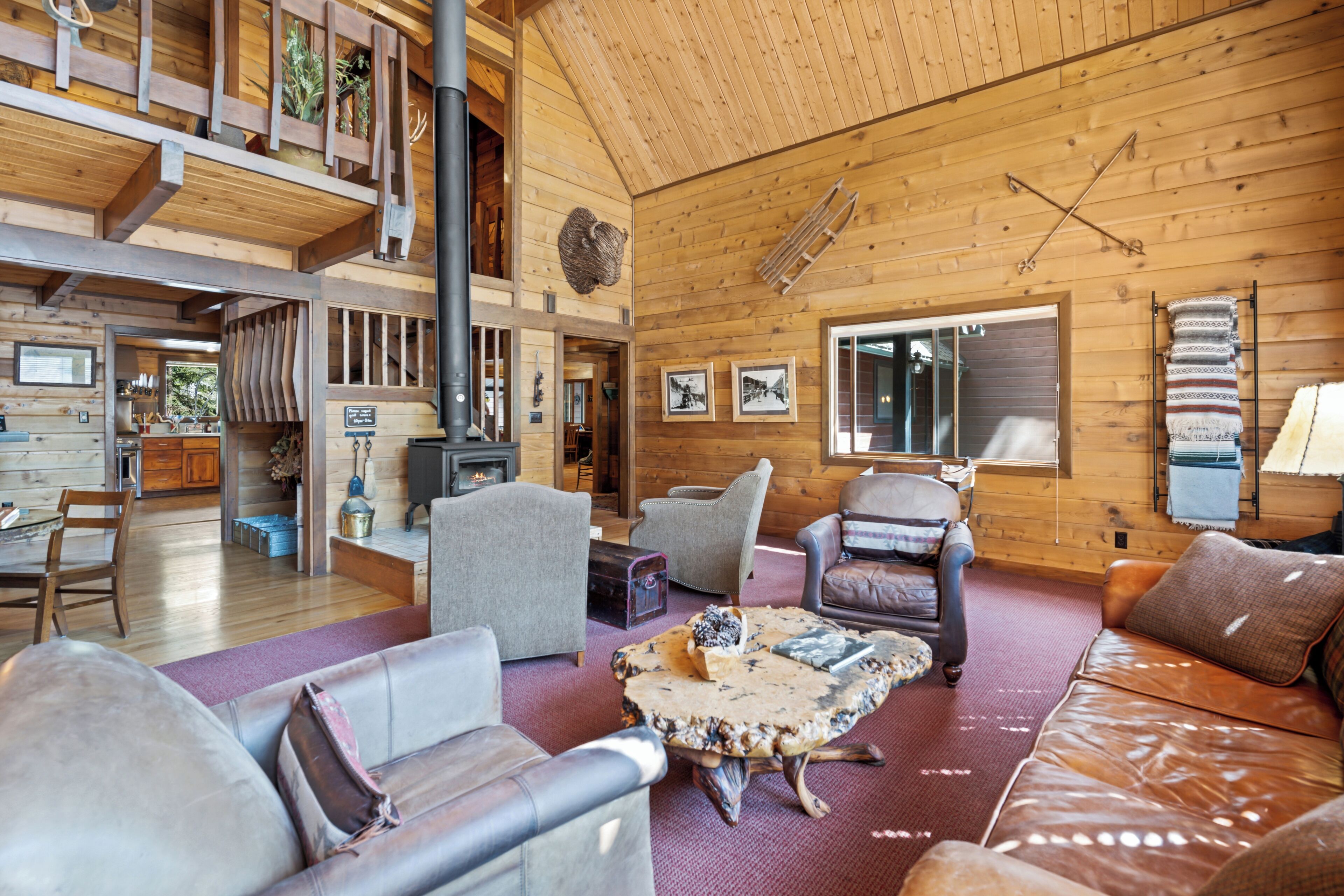 A spacious living area with a high ceiling, wooden walls, and a fireplace. There are multiple seating options including a sofa, chairs, and a coffee table.